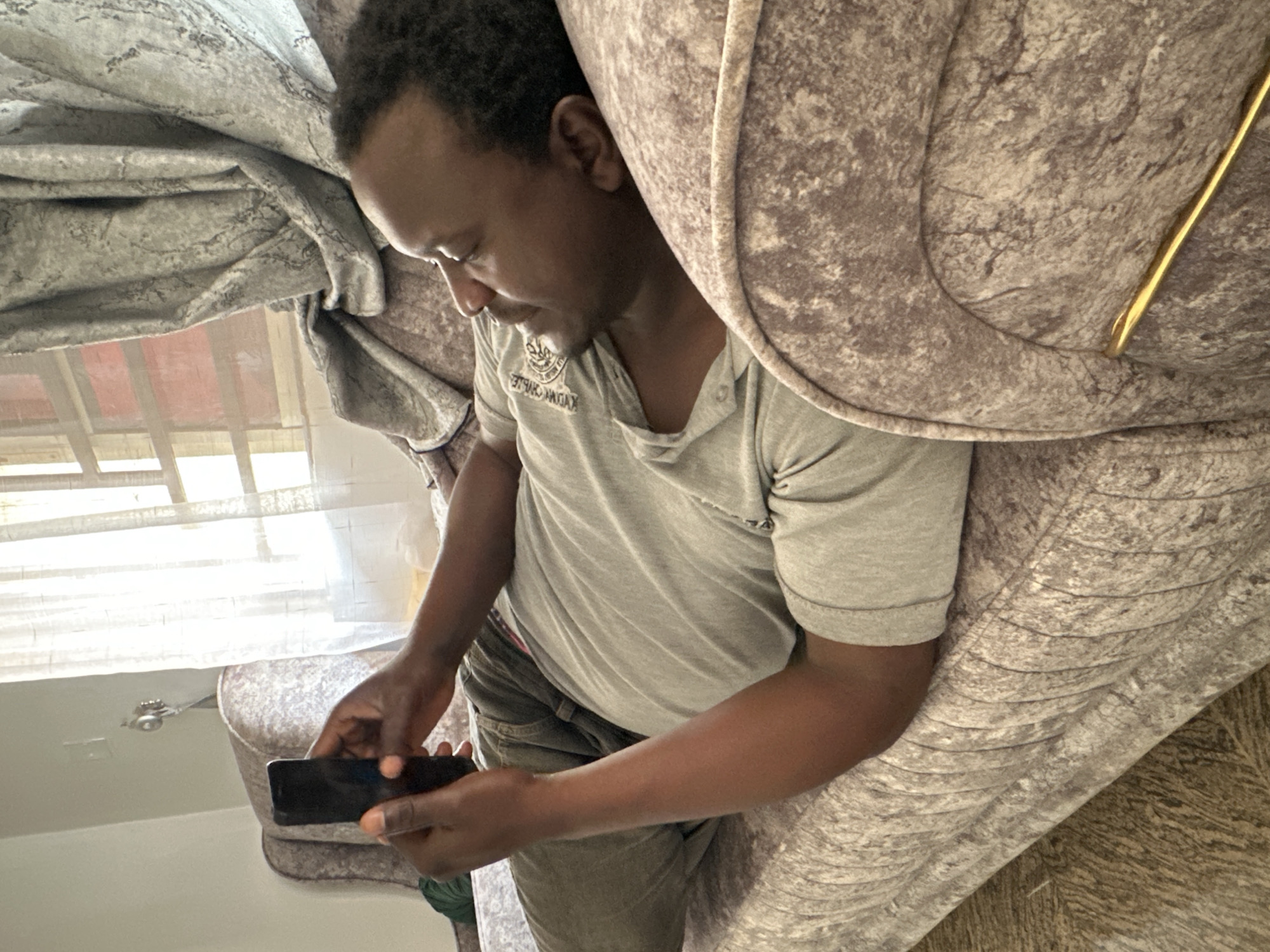 A man of African descent is lying on a gray, textured sofa, holding a black smartphone in his hands, and looking at it. He is wearing a gray t-shirt and dark pants. The room appears to be a living room, with a window and curtains visible in the background. The lighting suggests it is daytime, with natural light coming from the window. The man's posture indicates relaxation or engagement with the device. The overall environment is domestic and casual.