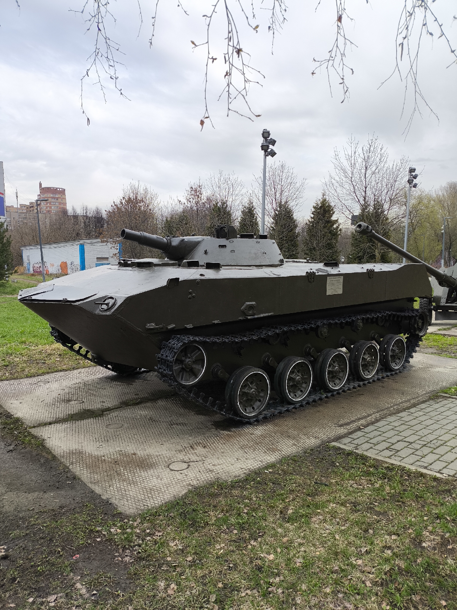 A wide shot captured on an overcast day features a dark olive green tracked military vehicle on static display. The vehicle, likely an Infantry Fighting Vehicle (IFV) or Armored Personnel Carrier (APC), has a low profile, a heavily sloped front hull, and a small turret equipped with a cannon. Its tracks and six road wheels are clearly visible. The vehicle rests on a patterned concrete slab, surrounded by patches of grass and dirt with fallen leaves.
In the background, bare tree branches hang from above, framing the scene. Behind the main vehicle, several leafless trees and some evergreen trees are visible, along with multiple streetlights or floodlights. To the right, part of another, larger military vehicle (possibly a self-propelled gun or tank) can be seen. Further in the distance, urban buildings are present, including a multi-story reddish-brown residential building and a smaller white building with graffiti.
A white rectangular plaque is affixed to the side of the main tracked vehicle. The visible Cyrillic text clearly reads "БМП-1" (BMP-1) and "Боевая машина пехоты" (Boyevaya Mashina Pekhoty), confirming it is a BMP-1 Infantry Fighting Vehicle. The overall setting suggests a military museum, park, or memorial site.