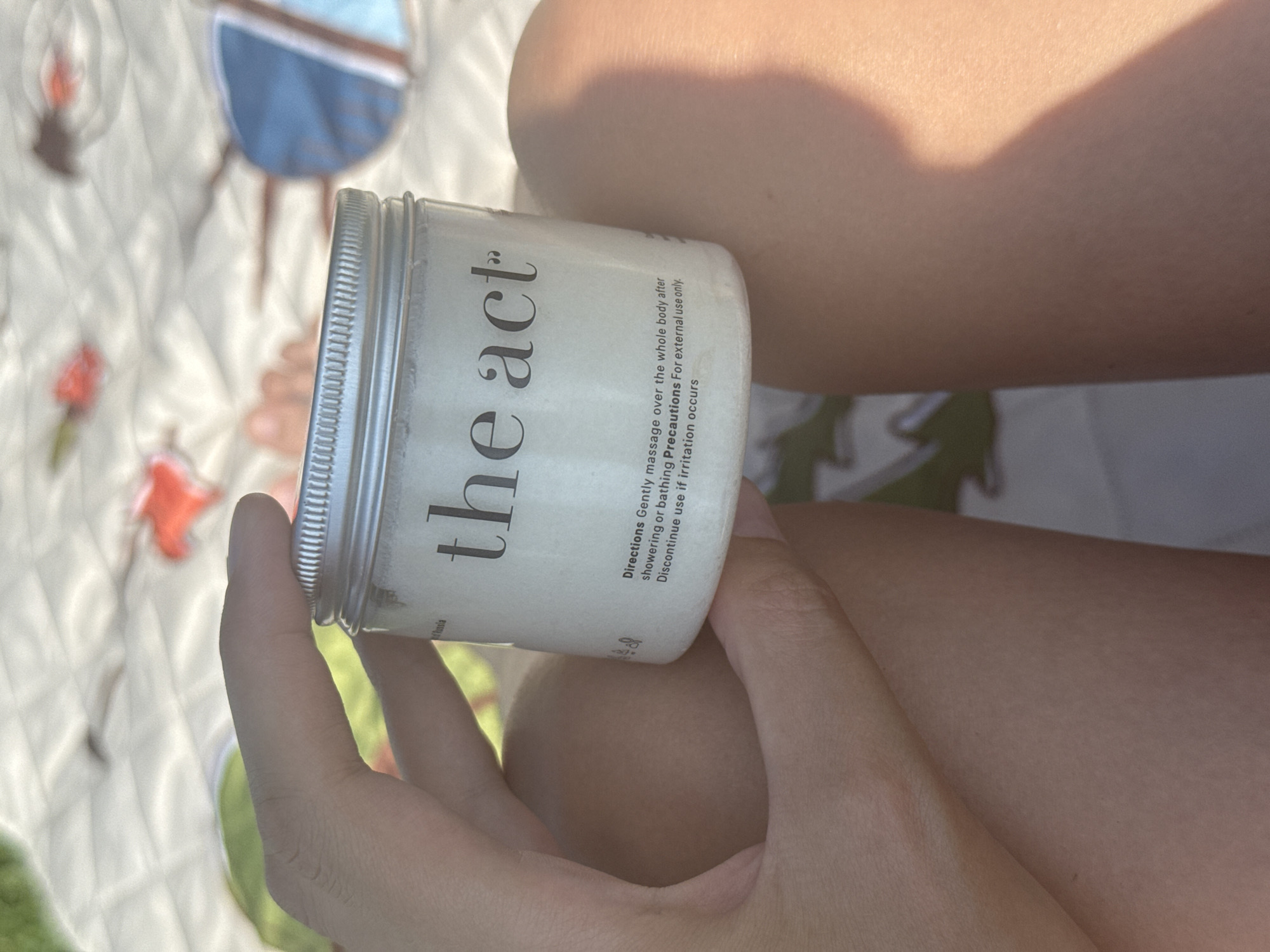 A person's hand holds a white jar with a silver lid. The label on the jar reads "the act". Below this, instructions are visible: "Directions Gently massage over the whole body after showering or bathing Precautions For external use only. Discontinue use if irritation occurs". The background appears to be a blanket with a floral pattern and a blurred image of a blue and white object. The scene is outdoors, likely in a park or at a picnic, with sunlight casting shadows. There is no indication of the city of Подшивалово, Russia, or any specific urban features.