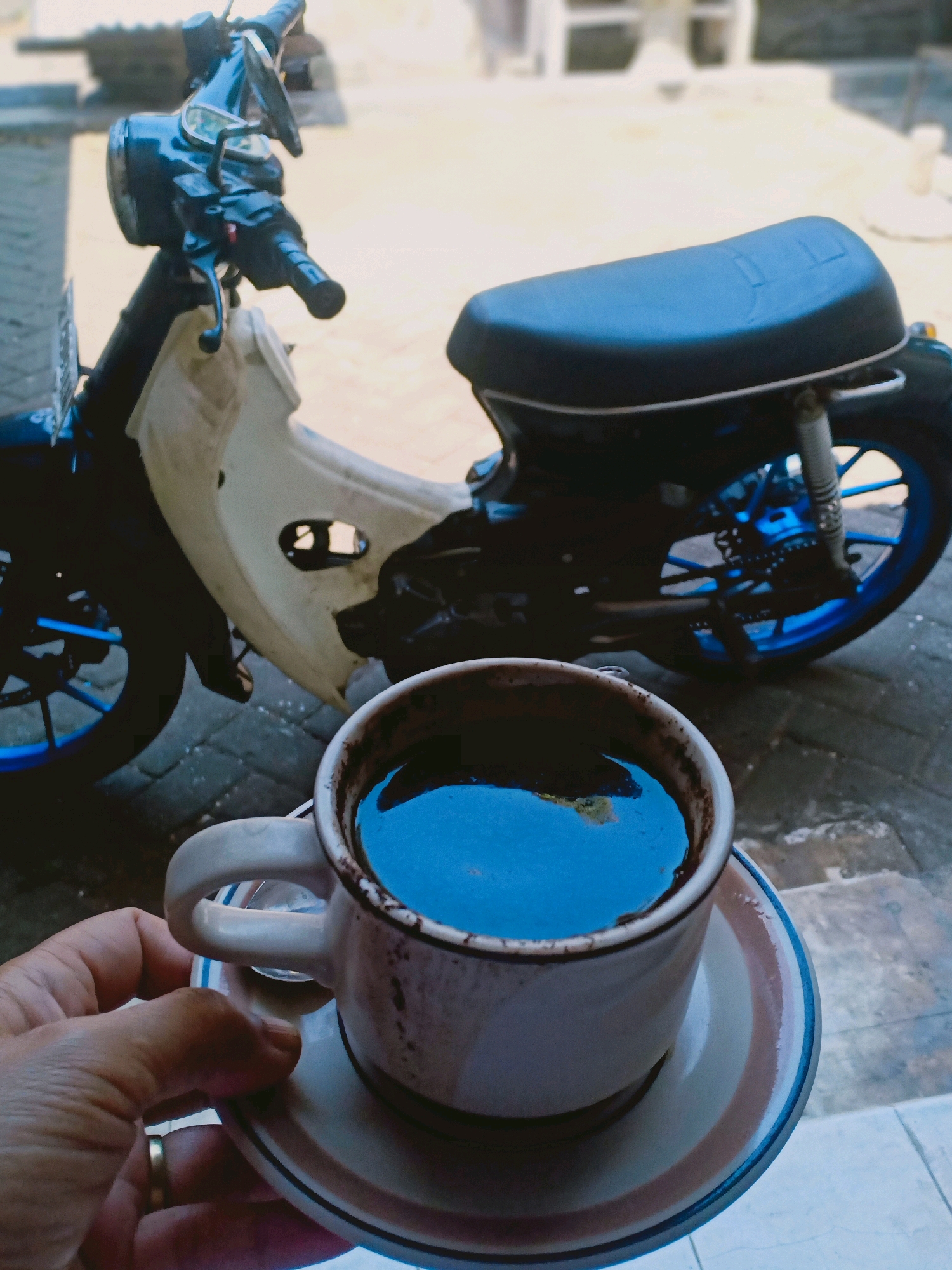 A hand holds a white mug filled with dark coffee, resting on a saucer. The mug and saucer have a striped pattern around the rim. The coffee's surface reflects the surroundings. In the background, a motorcycle is parked. The motorcycle is black and white with blue accents on the wheels. The scene appears to be outdoors on a paved surface, possibly a sidewalk or driveway, with hints of a building and possibly a road in the blurred background. The lighting suggests it is daytime. The location is identified as Lumajang, Indonesia.