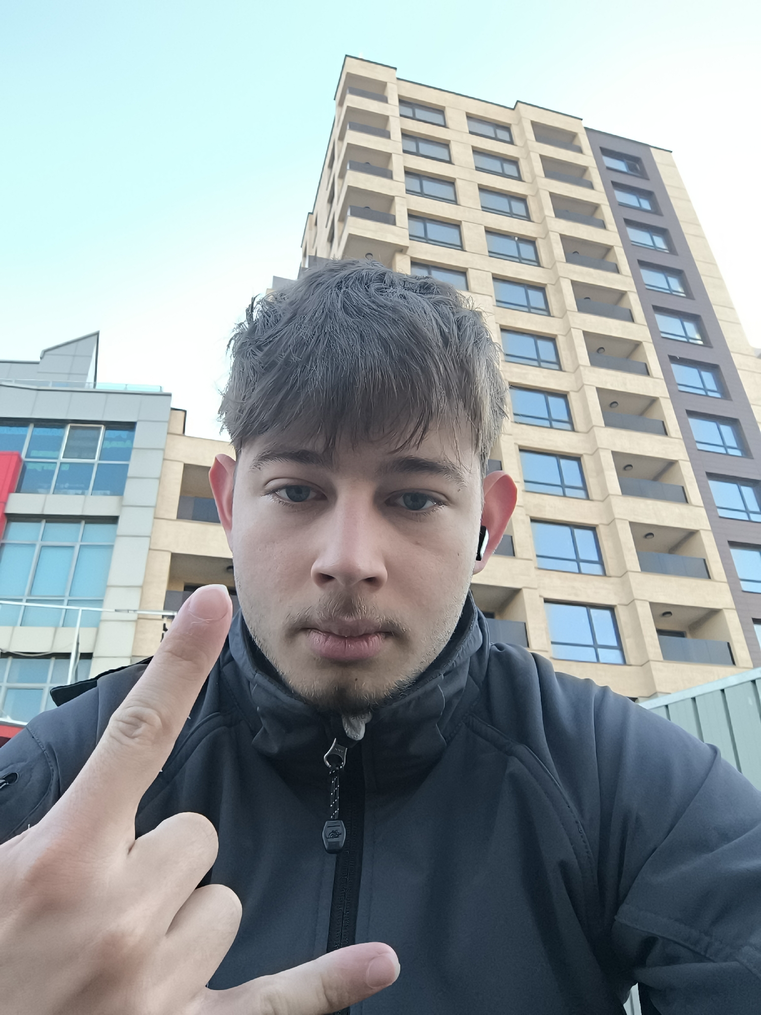 A selfie shows a light-skinned, young male wearing a black jacket with a zipper pull tag and a single wireless earbud in his right ear. His hair is dark, and he has a light beard. He is holding up his index finger, while the rest of his fingers are bent back. Behind him are tall buildings, one taller than the other. The taller building is light brown and has multiple rows of windows and balconies. The shorter building is a modern architectural style with blue windows and a red accent. The sky is pale blue. The photograph appears to have been taken outdoors in daylight. While not definitively clear, the architectural style of the buildings in the background suggests the image could be from the city of Sofia, Bulgaria, though this cannot be confirmed without further context.