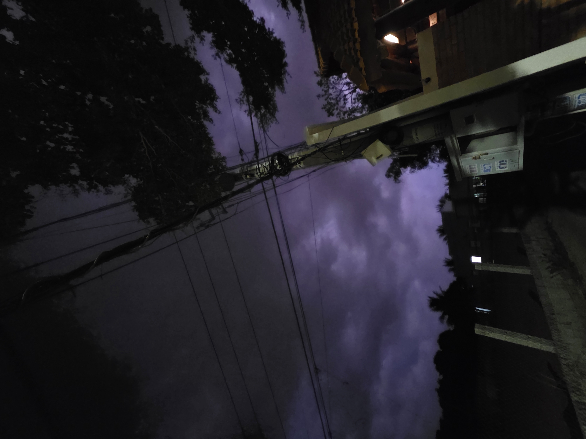 The image, taken at night in Mũi Né, Vietnam, shows a low-angle view towards the sky, filled with dark, stormy clouds. Power lines crisscross the view. Street lights illuminate a building on the right side, made of beige brick and concrete. Trees are visible on the left and right.