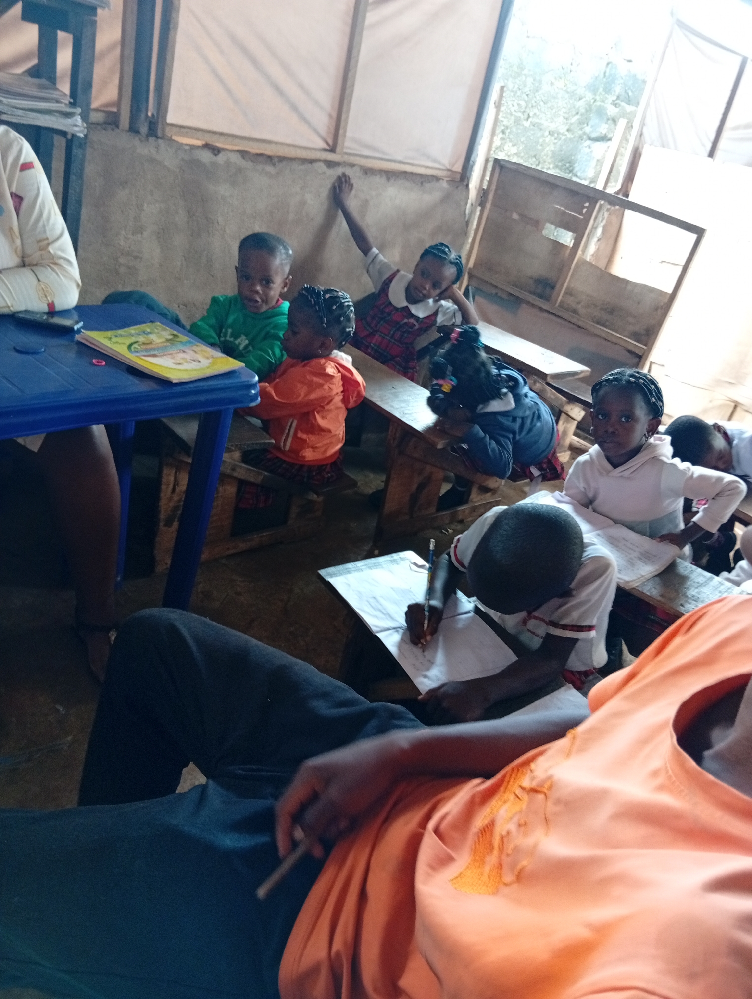 In a classroom setting in Unknown, Unknown, several children are seated at wooden desks. The desks are arranged in rows, with the children facing forward. Some children are writing on paper, while others are looking towards the front of the room. A teacher is seated at a blue table in the foreground, with a book on the table. The classroom appears to be sparsely furnished, with simple wooden frames visible.