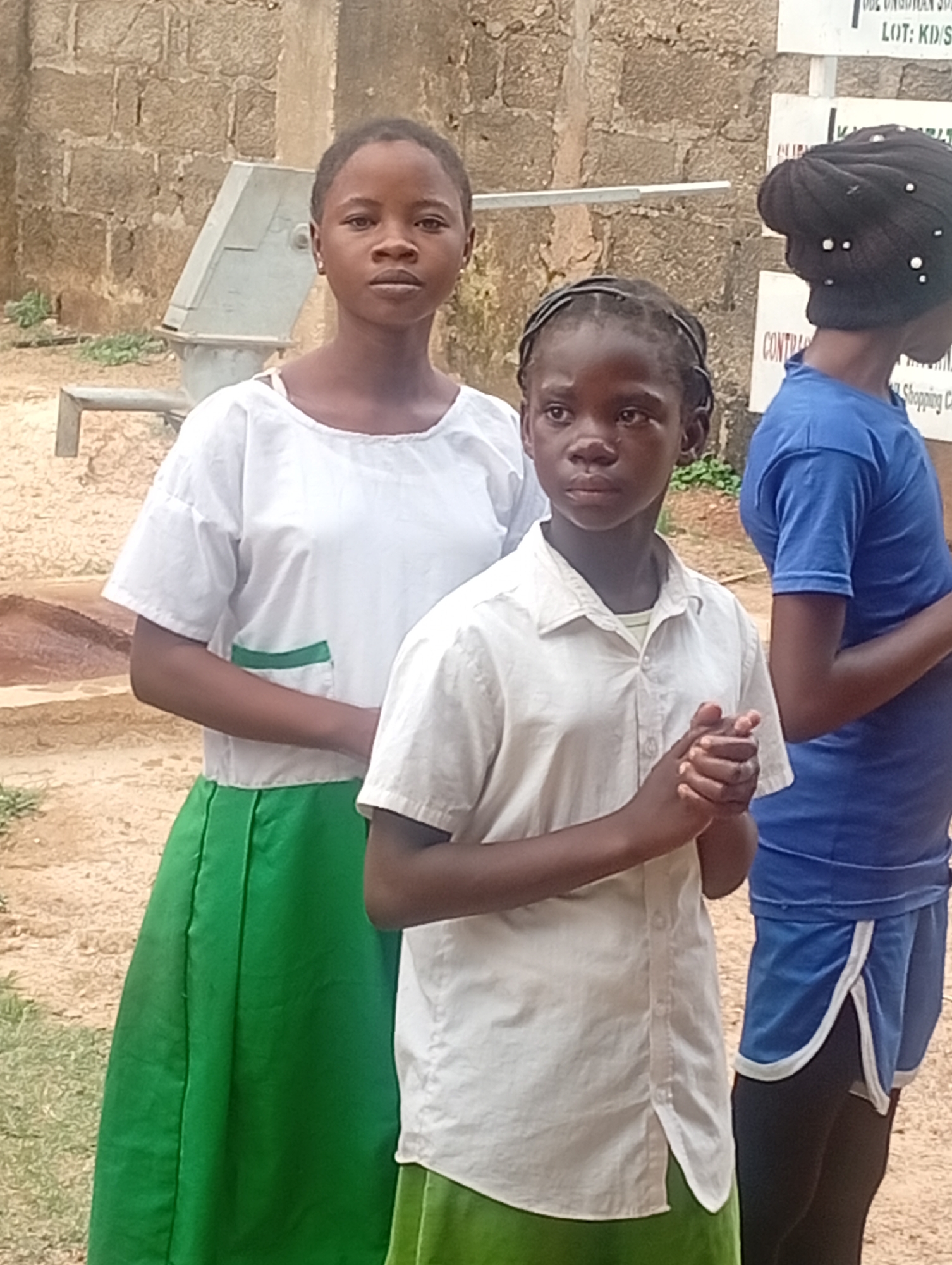 Three young individuals are the focal point of the image. Two are situated towards the left side, while the third is partially obscured on the right. The individuals appear to be positioned outdoors in an urban environment, as indicated by the presence of a brick wall and a mechanical apparatus in the background, along with a sign with text.
The first individual on the left is attired in a white top, distinguished by a green pocket, and a green skirt. The second individual is dressed in a white shirt and a green skirt. The third, positioned on the right, wears a blue top, blue shorts with a white trim, and a black head covering. The second individual is holding their hands together in front of them.
The scene is illuminated by natural daylight, suggesting an outdoor setting. Additional components within the frame include the brick wall, the mechanical apparatus, and the sign with legible text. The locale is identified as Unknown, Unknown.