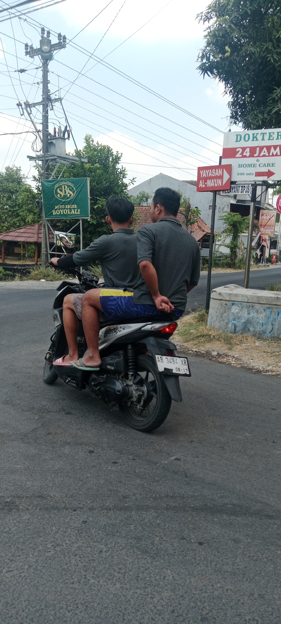 Two men ride on a single scooter on a paved road. The rider in front, wearing a grey polo shirt and patterned shorts, is looking forward and appears to be driving. The passenger behind him, also in a grey polo shirt, has his arms around the rider in front. Both men are casually dressed, with the passenger wearing blue and yellow patterned shorts. The scooter is a modern, dark-colored model with a license plate visible that reads "AB 3494 XR". In the background, a green sign reads "SOTO SEGER BOYOLALI" and a red and white sign indicates "DOKTER 24 JAM & HOME CARE". Trees and buildings line the road, and power lines are visible overhead. The scene appears to be in a semi-urban or rural area in Maguwoharjo, Indonesia, given the style of the signs and the general environment.