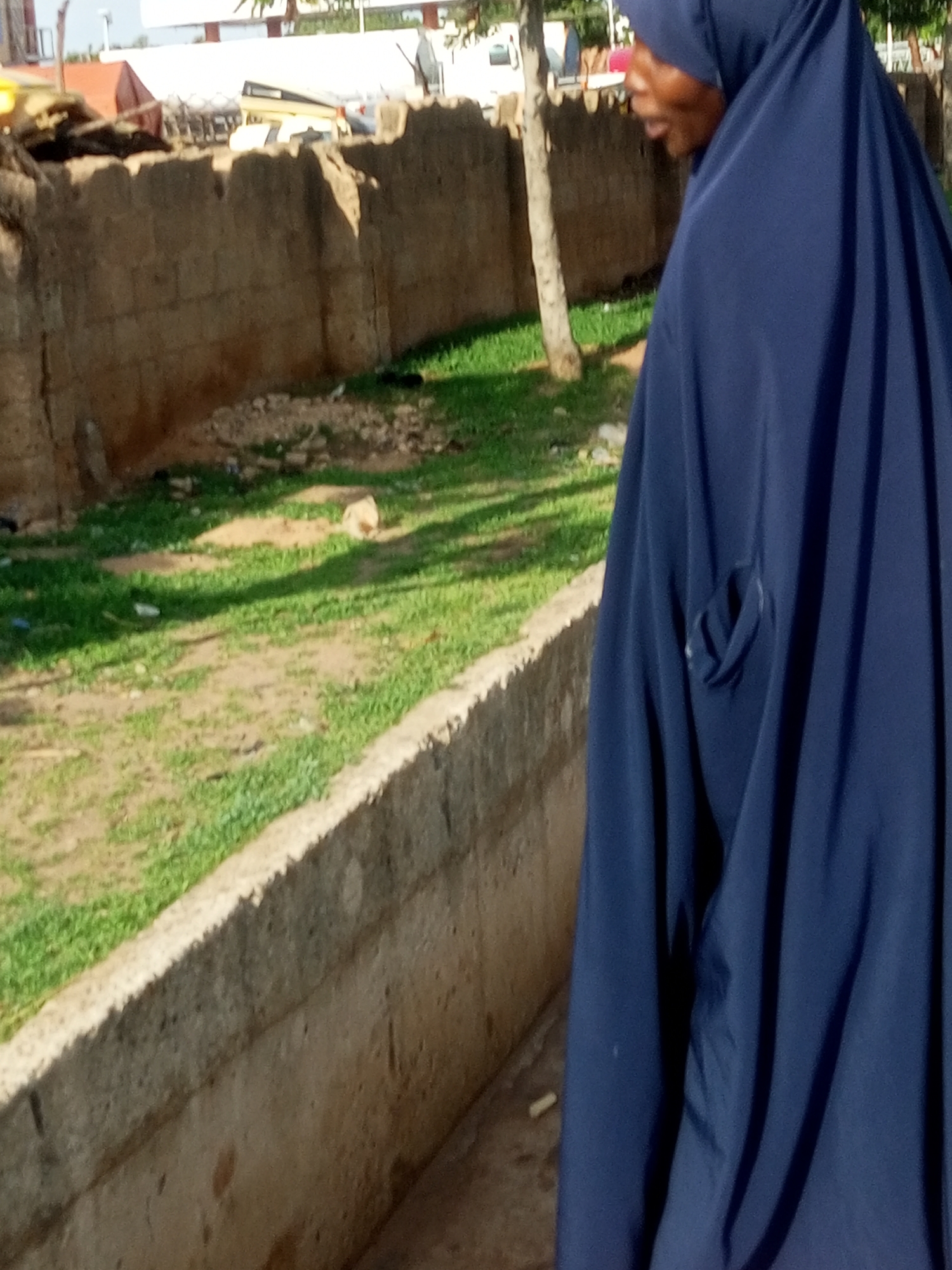 The image shows a person wearing a dark blue hijab walking outdoors. The person is partially visible, with their face turned to the side and their body angled away from the camera. The hijab covers their head and body, with folds and creases creating a sense of depth. A dark pocket or flap is visible on the side of the garment.
The background suggests an outdoor setting with a stone wall running along the left side of the frame. Beyond the wall, there is a patch of green grass, with some dirt and debris scattered on the ground. A tree trunk is visible behind the person, and further in the distance, there are hints of buildings and structures, possibly under construction or in a state of disrepair. The lighting suggests it is daytime, with sunlight casting shadows on the grass.
The overall impression is one of a casual, everyday scene in an urban or semi-urban environment. There are no clear indicators of a specific event or strong emotions, beyond the implied movement of the person. The location context provided as Gombe, Nigeria, suggests this scene is set in that region. No visible text is present in the image.