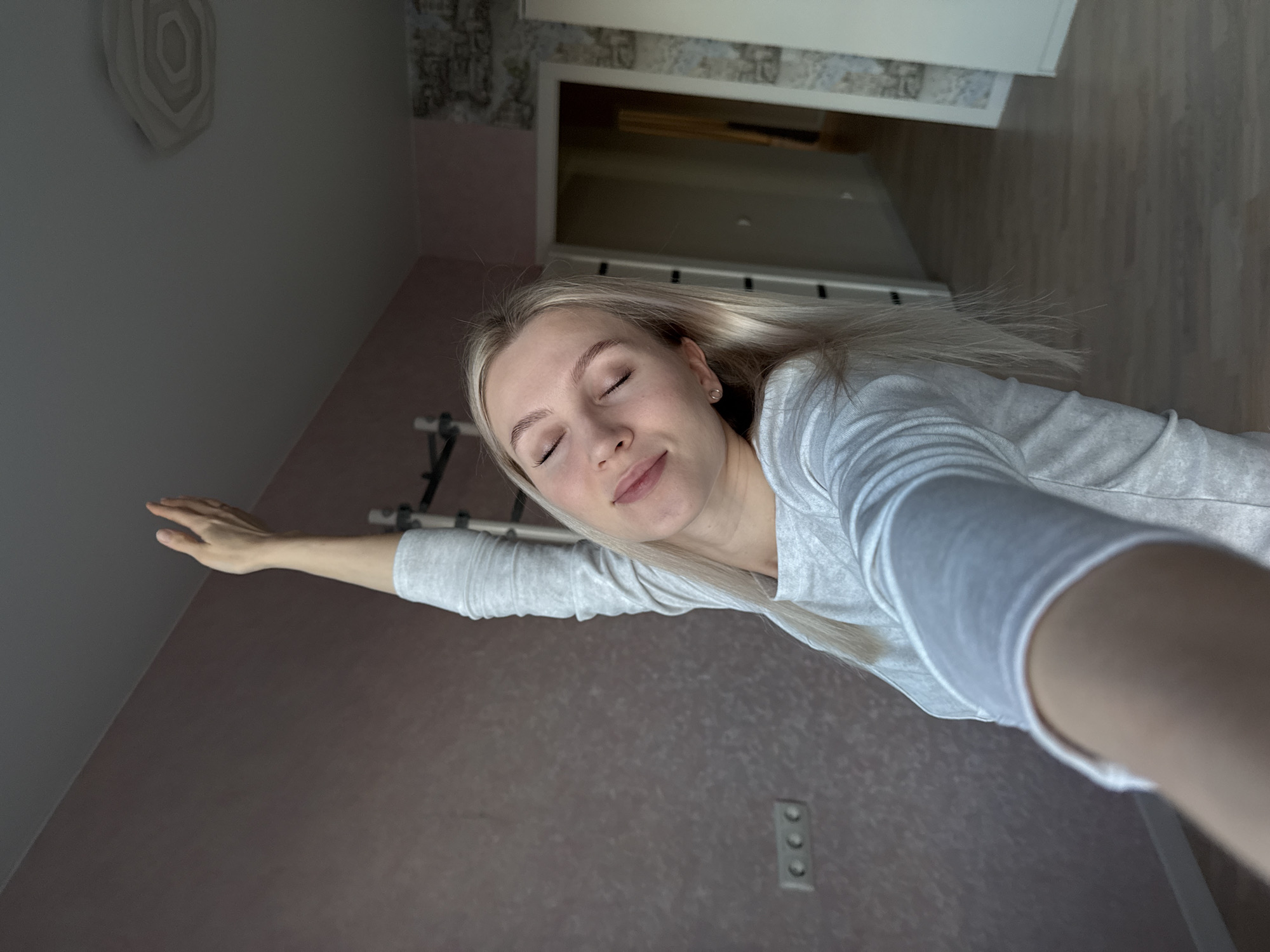 A woman with blonde hair is lying down, eyes closed, and smiling slightly. She is wearing a gray long-sleeved shirt. Her right arm is extended to the left of the frame. The location appears to be indoors, possibly a room with pale gray and maroon walls. There is a wall decoration above her head. In the background, there is a doorway leading to another room with a light-colored wooden floor. An electrical outlet is visible on the maroon wall. The image gives the impression of a self-portrait taken from an unusual angle. There's no definitive evidence to place the image in Izhevsk, Russia, so that cannot be verified.