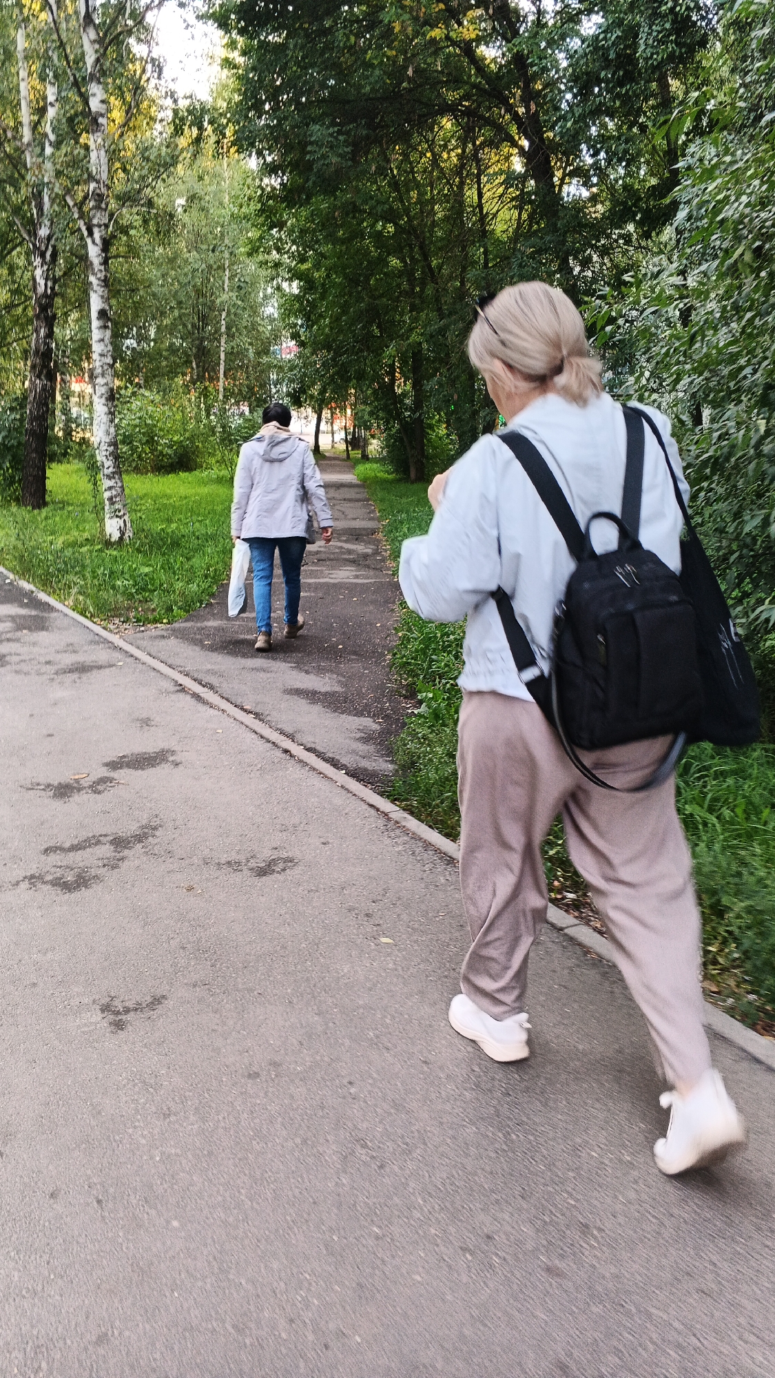 Two individuals are walking on a paved path in a park-like setting, likely in Izhevsk, Russia, given the context. The person in the foreground, facing away from the camera, is a woman with blonde hair pulled back, wearing a light blue jacket, tan pants, and white sneakers. She has a black backpack with a white logo on her back. The person further down the path, also walking away, appears to be a woman wearing a light gray hooded jacket, blue jeans, and brown shoes, carrying a white plastic bag in her right hand. The path is bordered by a grassy area on the left and dense green foliage on the right. Trees with white bark and green leaves are visible on the left side of the path. The lighting suggests it is daytime, possibly late afternoon or early evening, with dappled sunlight filtering through the trees. The overall scene depicts a casual outdoor stroll in a city park.