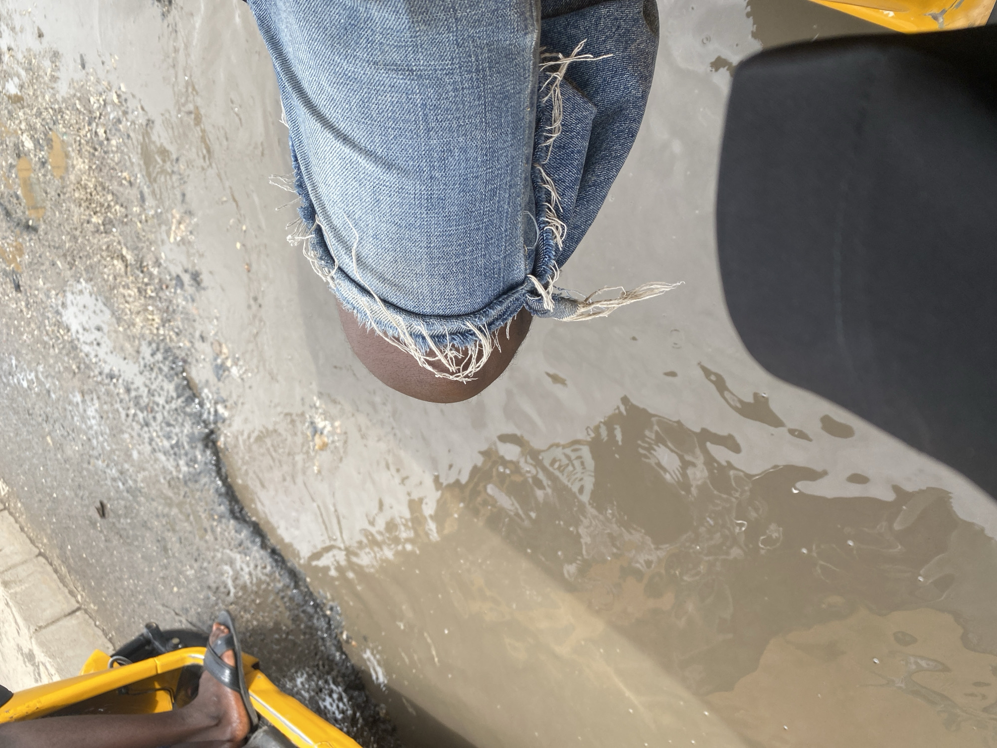 This image shows a close-up view of a person's legs and feet, dressed in blue jeans and black sandals. The person is seated in what appears to be a yellow vehicle, possibly a tricycle or tuk-tuk, as indicated by a yellow frame and a black seat. The scene depicts a flooded area with muddy water on the ground, suggesting recent rain or poor drainage. The ground is a mix of asphalt, gravel, and water, with some debris visible. The jeans have a distressed, frayed hem. The lighting suggests it is daytime. The location is identified as Jalingo, Nigeria, and the overall impression is of a functional, everyday scene in a tropical or sub-tropical urban environment, likely during or after a period of rain. No text is visible in the image.
