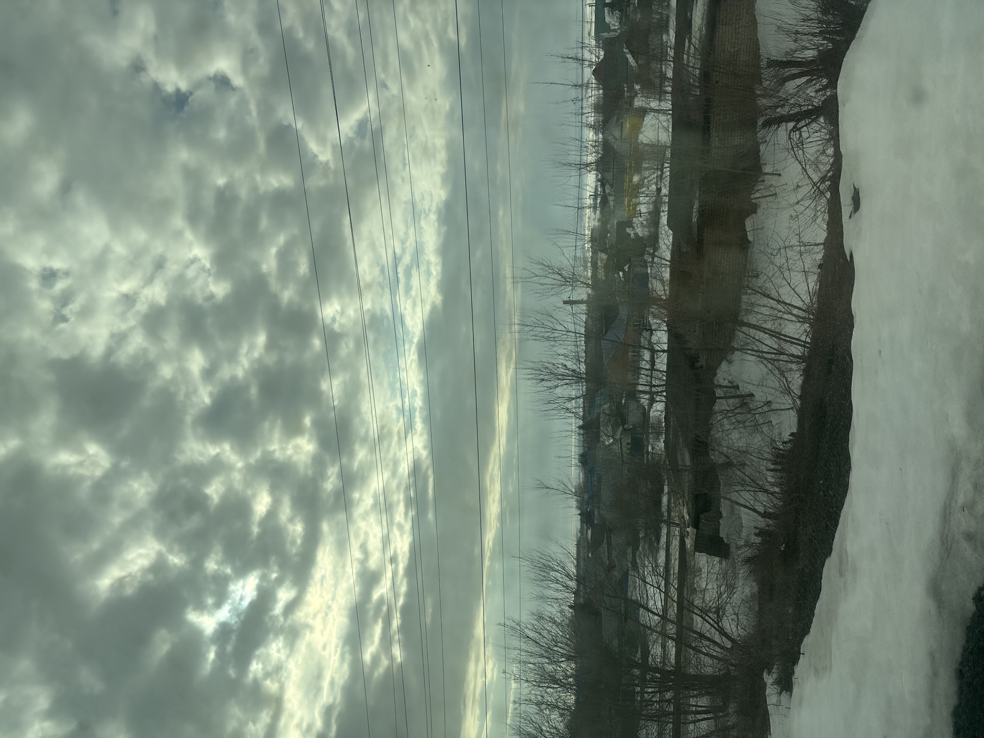 The image presents an elevated view of a winter or early spring landscape in Nurlat, Russia, under a heavily overcast sky featuring textured, grey-white clouds with brighter patches indicating obscured sunlight in the upper left. Multiple dark, horizontal utility lines traverse the upper-mid section of the frame. Below these lines, the distant horizon reveals a flat, snow-covered expanse, possibly a frozen body of water or field, under a lighter sky. The midground contains a sparse residential settlement with various small buildings, identifiable by their roofs in shades of dark green, dark red, brown, and grey. Bare-branched deciduous trees and dense shrubs are scattered throughout the settlement and in the foreground. A singular, slender, vertical structure, possibly a chimney or vent, is visible above the houses. The ground is largely covered in snow, with areas of dark, exposed earth and vegetation, particularly prominent in the foreground. No people, specific actions, or interactions are visible within the scene.