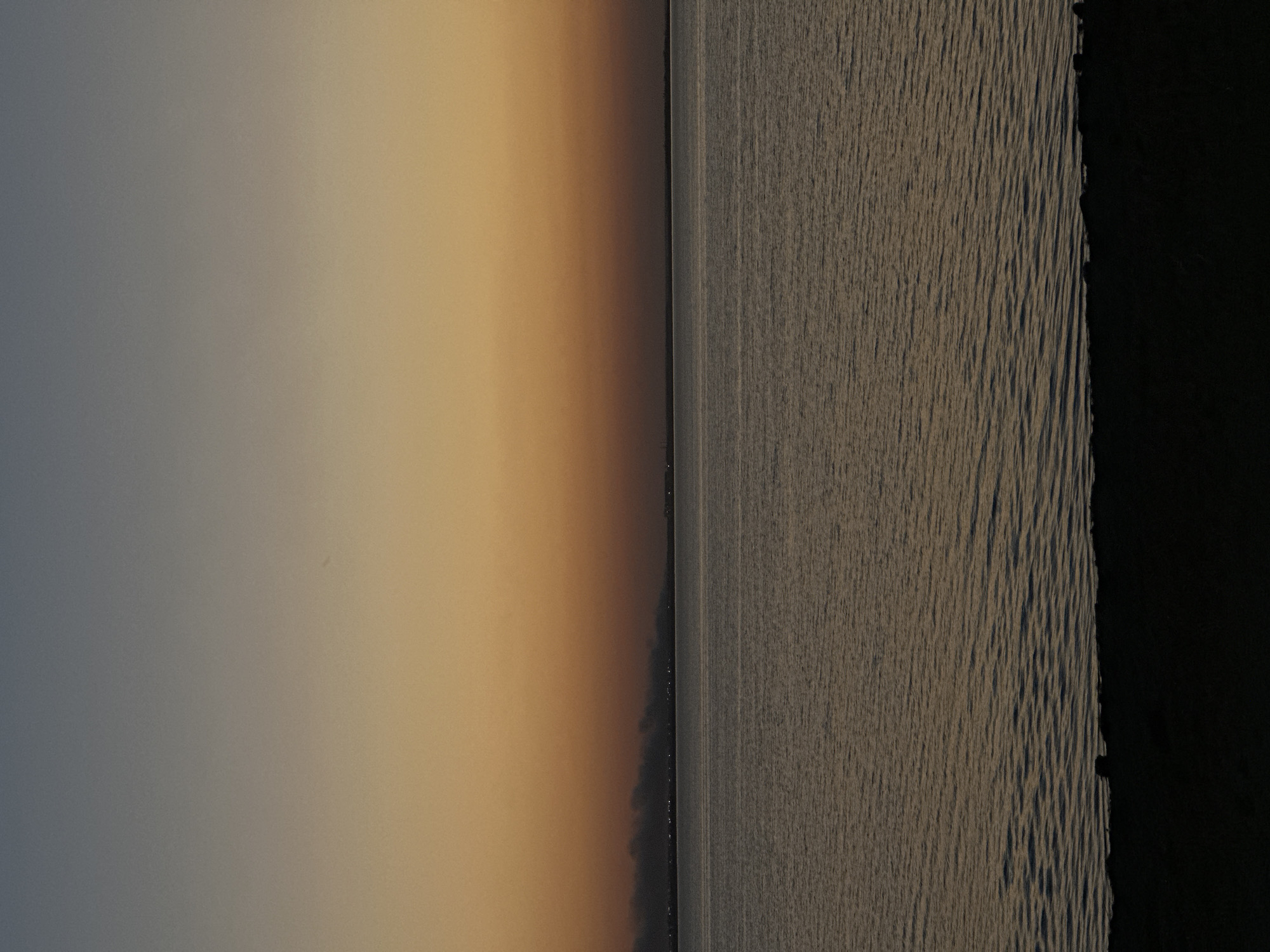 This is a vertically oriented, low-angle, full-shot photograph of a body of water under a twilight sky. The sky transitions from a pale blue at the top to a warm orange near the horizon. Along the distant horizon, faint lights and plumes of smoke are visible, suggesting an urban or industrial area. The water in the foreground is calm with subtle ripples reflecting the muted colors of the sky. The bottom of the frame is occupied by a dark, silhouetted shoreline. The overall scene evokes a sense of quietude and dusk. The image depicts the Big Kokshetau Lake in Kokshetau, Kazakhstan, at the time of sunset.