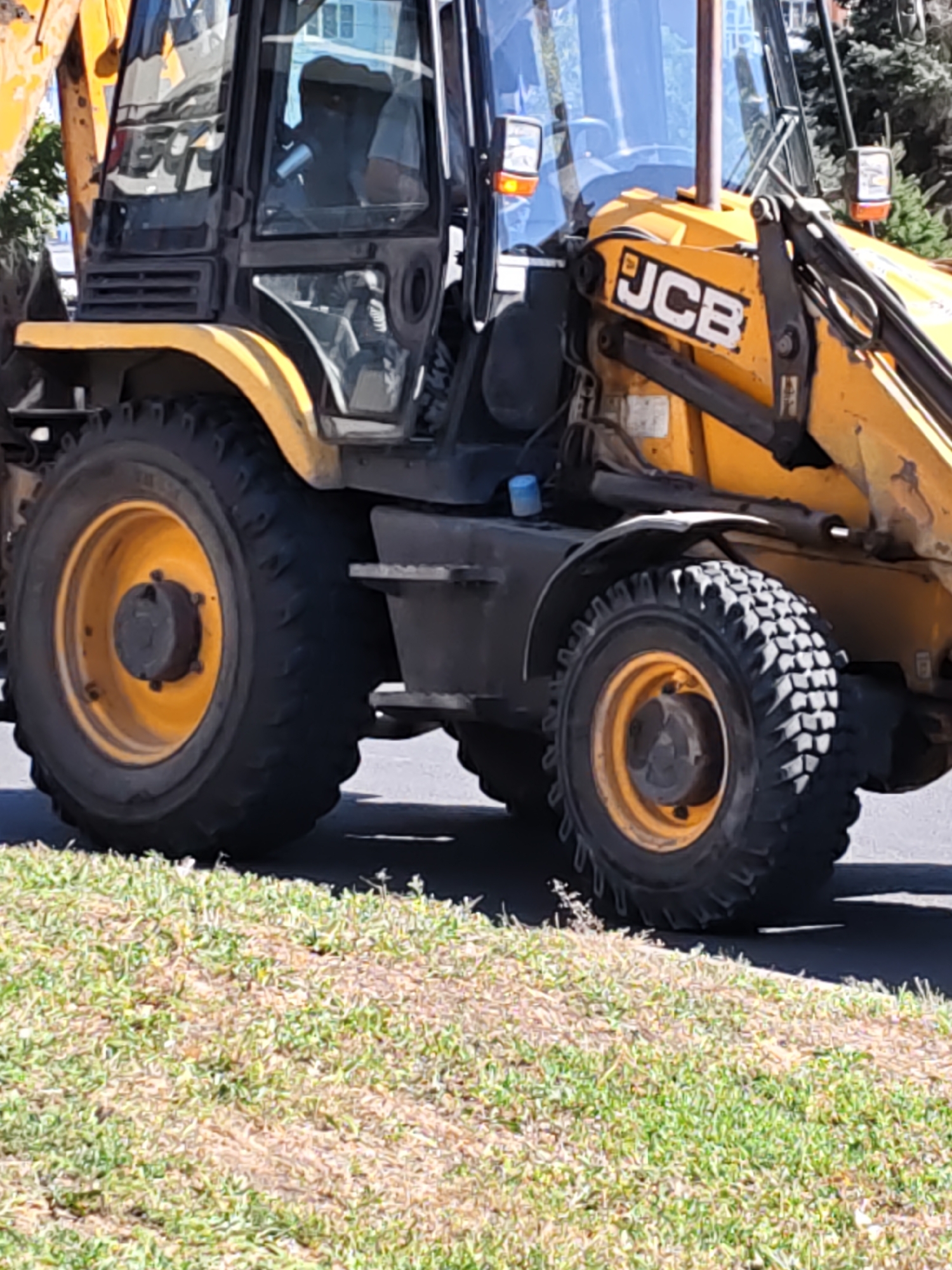 The image shows a yellow JCB backhoe loader parked on a paved surface next to a grassy area. The backhoe has large, thick tires with a distinctive tread pattern. The front of the machine is visible, including the cab with tinted windows. A partial view of the JCB logo is present on the side of the machine. The scene appears to be outdoors, possibly in an urban or suburban setting, given the presence of pavement and the hints of buildings in the background reflected in the windows. There are no people clearly visible in the image, though figures are faintly discernible within the cab. The lighting suggests it is daytime. The location is identified as Rostov-on-Don, Russia.