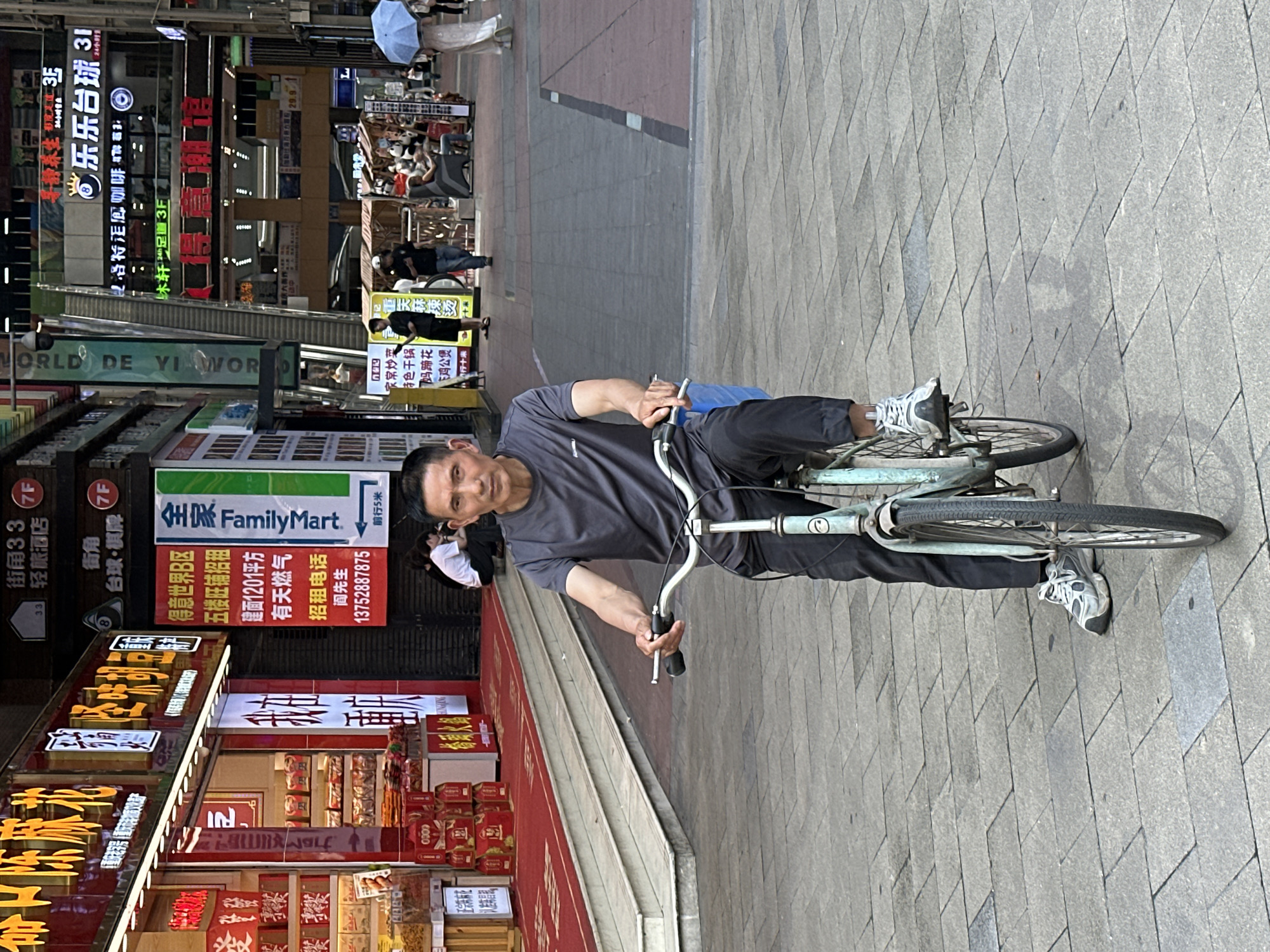 A man is riding a bicycle on a brick-paved sidewalk. He is wearing a gray shirt and black pants, and he is looking at the camera. In the background, there is a street with shops and signs, including a FamilyMart and signs in Chinese characters. The street is in the Yuzhong District of Chongqing, China.