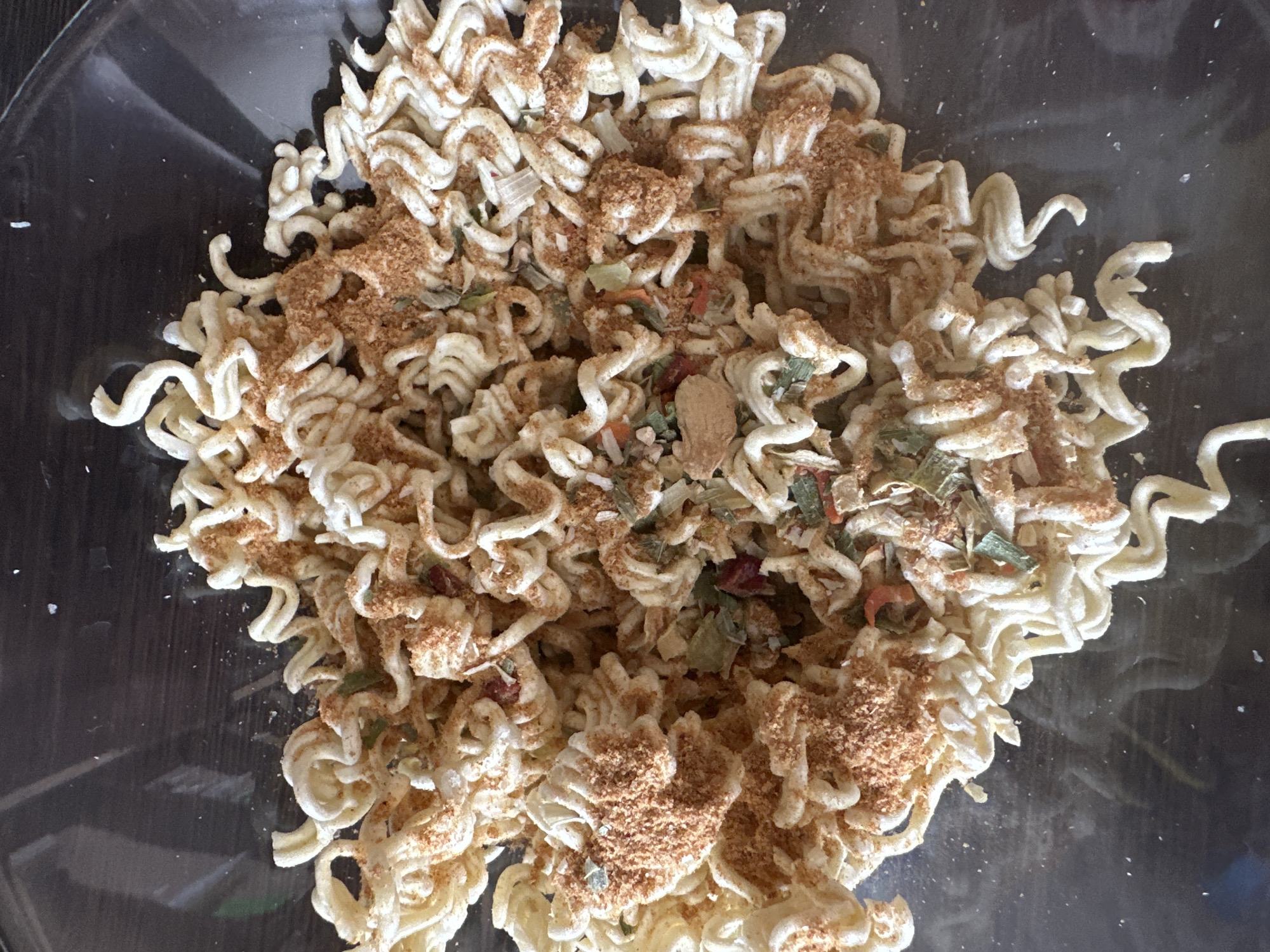 The image shows a close-up, top-down view of uncooked ramen noodles in a clear bowl. The noodles are dry, coiled, and appear to be seasoned with a reddish-brown powder and small flecks of green and red dehydrated vegetables. The bowl is placed on a dark, possibly wooden surface. There is no indication of people, actions, or interactions. The location in Novoaltaysk, Russia is not discernible from the image.