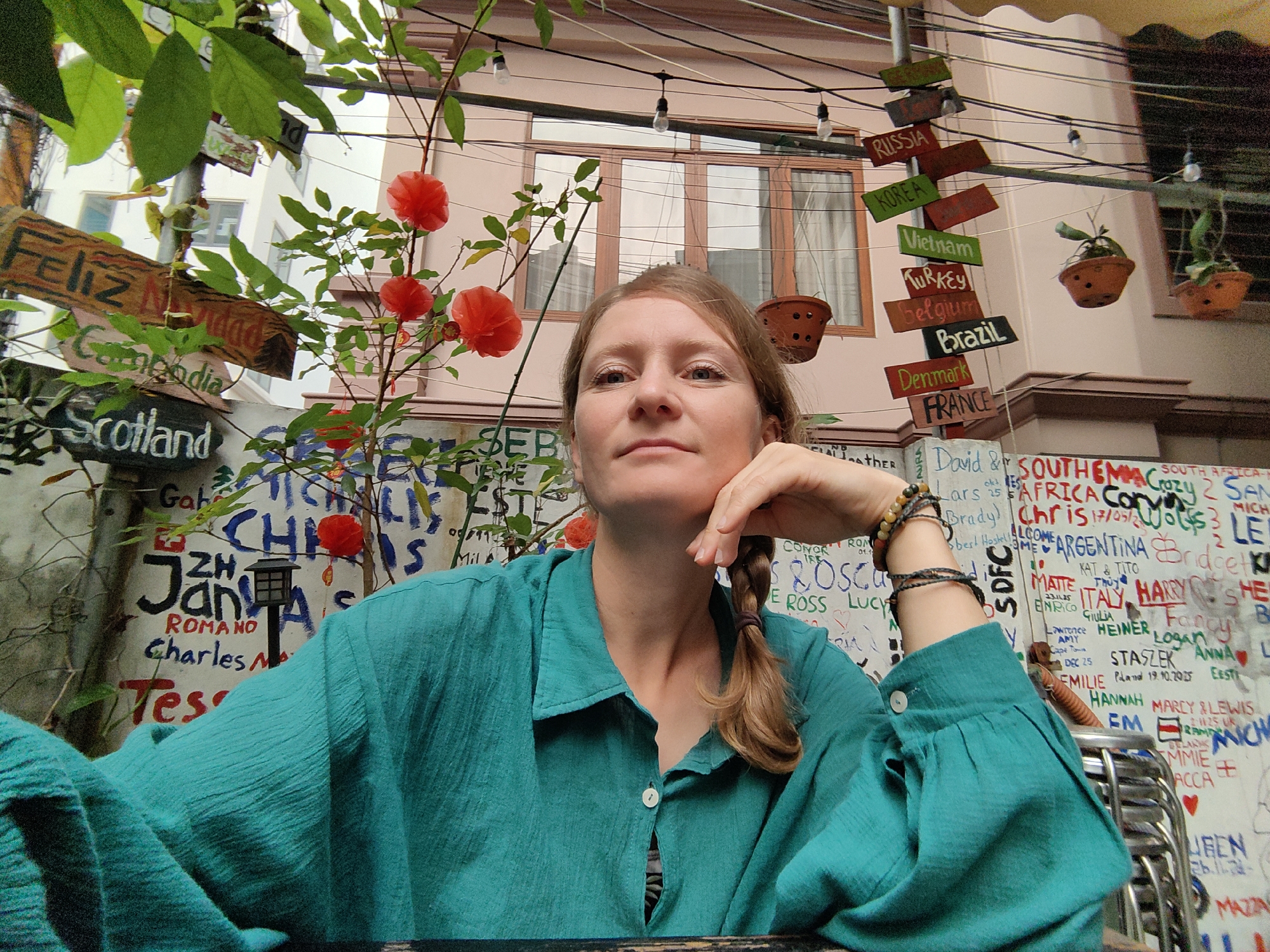 A medium shot shows a fair-skinned woman with light brown hair styled in a side braid, wearing a teal button-down shirt, seated at a table. She rests her chin on her hand, which is adorned with bracelets. Behind her is a wall covered in graffiti, including names and dates. To the upper right, signs pointing to countries like Russia, Korea, Vietnam, and Brazil hang from overhead wires. Potted plants are also suspended from the wires. A tree with bright red spherical decorations sits to the left. The location is identified by signage to be in Bai Chay Ward, Vietnam. The woman appears to be a customer at an outdoor cafe with graffiti walls in Bai Chay Ward, Vietnam.