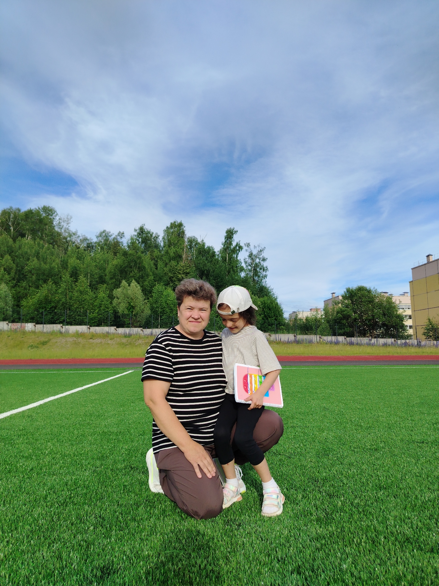 Two people are kneeling on a bright green artificial turf field, likely a sports field, with white lines visible. The person on the left is an adult woman with short, curly, brown hair, wearing a black and white striped t-shirt and brown pants. She is smiling at the camera. The person on the right is a young child, possibly a girl, wearing a white baseball cap, a striped t-shirt, black leggings, and white sneakers. She is looking down at a pink object, which appears to be a notebook or book, with colorful elements on its cover.
The background features a line of lush green trees and some buildings in the distance. The sky above is a bright blue with wispy white clouds. The overall scene suggests a bright, clear day. The presence of the sports field and the attire of the subjects suggest an outdoor recreational or educational activity. The people appear happy and relaxed, with the adult woman's smile directed towards the viewer and the child engrossed in her book. The location is identified as Noviy, Russia. No visible text is present in the image.