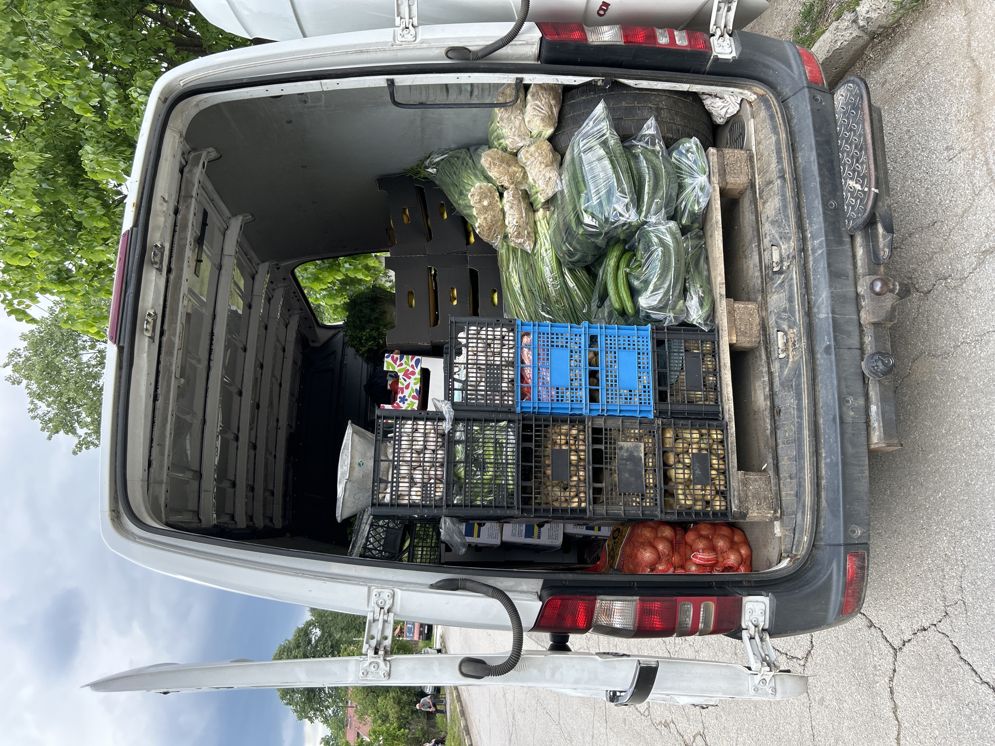 The image shows the open back of a white van, loaded with produce in crates and bags. The produce includes cucumbers, onions, potatoes, garlic, and dill. The van is parked on a cracked asphalt road. The location is listed as Unknown, Unknown.