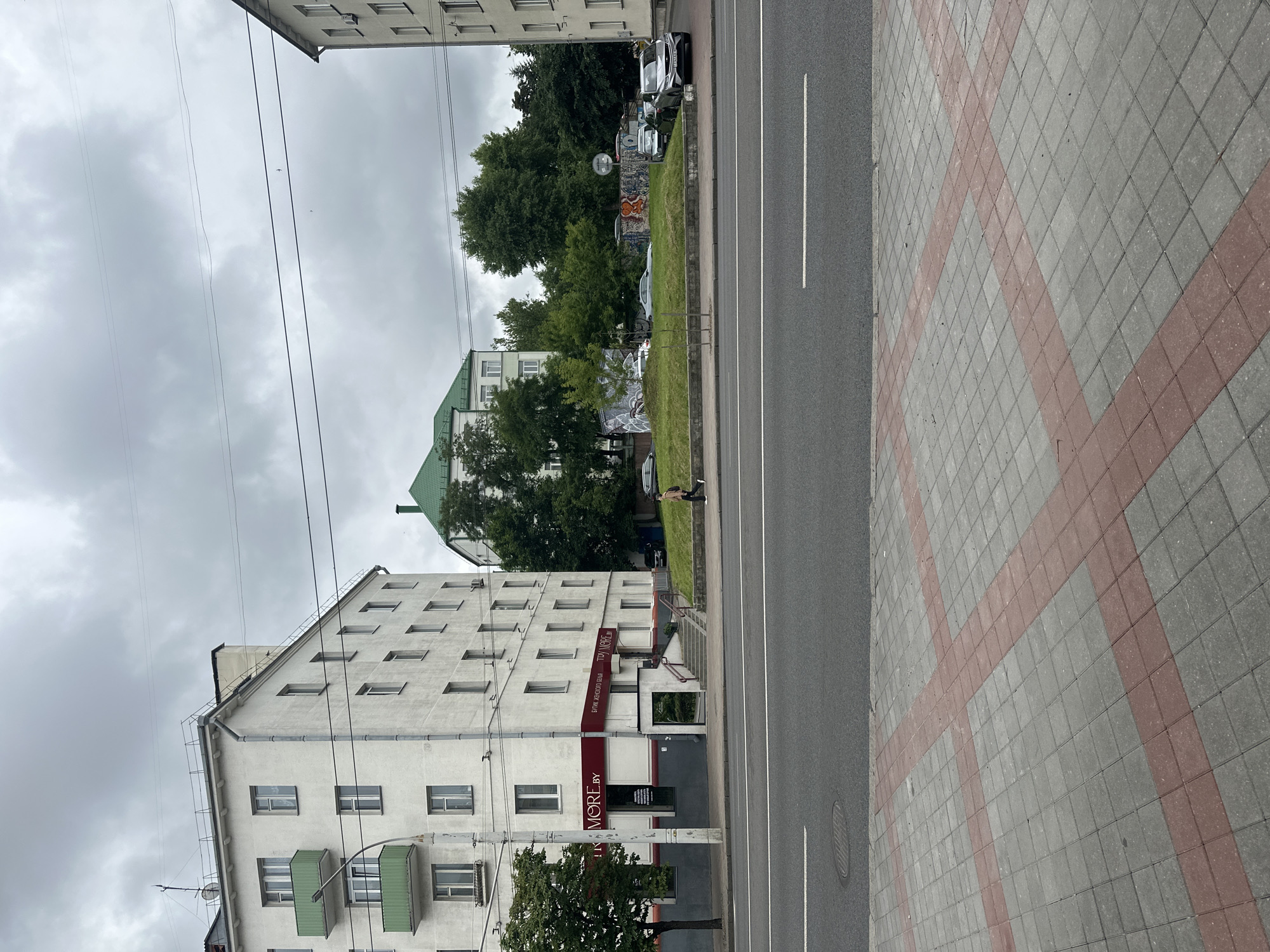 In Minsk, Belarus, on a cloudy day, a street scene unfolds with several buildings and a roadway. On the left, a white multi-story apartment building with green balconies features a business on the ground floor with a red sign that reads "MORE.BY" in English and Russian. Further back, another apartment building with a green roof is visible, nestled among trees. The street in the foreground is paved with asphalt and bordered by a sidewalk with red and gray interlocking pavers. A lone pedestrian walks on the sidewalk, heading away from the camera. Cars are parked on the grass embankment behind the sidewalk, and some graffiti adorns a low wall in that area. The sky is overcast, and power lines are visible crisscrossing the frame.