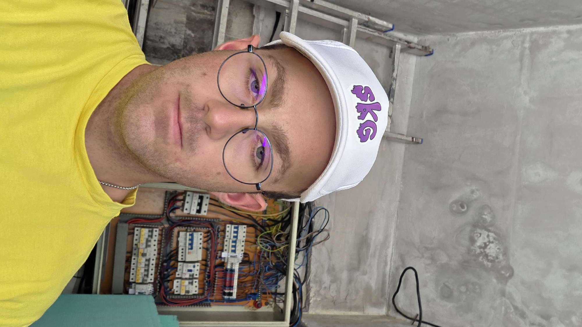 A medium shot shows a fair-skinned man wearing a white baseball cap with a purple "SKG" logo and round, wire-rimmed glasses. He has short brown hair and is wearing a bright yellow t-shirt with a silver chain necklace. The man is looking directly at the camera. In the background, to the right of the man, is an electrical panel with numerous wires and circuit breakers. To the left of the man is a silver ladder. The walls appear to be concrete and unfinished. The scene is indoors in what seems to be a construction or renovation site.