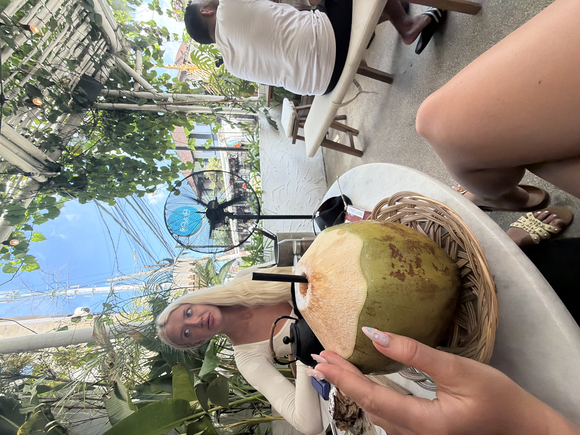 The image depicts a woman with blonde hair seated at a table outdoors, possibly at a cafe in North Kuta, Indonesia. She is wearing a white long-sleeved top and looking to her left. In front of her on the table is a whole coconut with a straw, a black teapot, and a plate with a slice of dessert. A hand with manicured nails is reaching towards the coconut. In the background, a man is seated on a bench, wearing a white t-shirt and black shorts. The environment appears to be lush with greenery, including large potted plants and vines hanging from a white, slatted roof structure. A large black fan is visible behind the woman. The lighting suggests it is daytime, with clear blue skies visible above. The overall scene conveys a relaxed, tropical ambiance.
