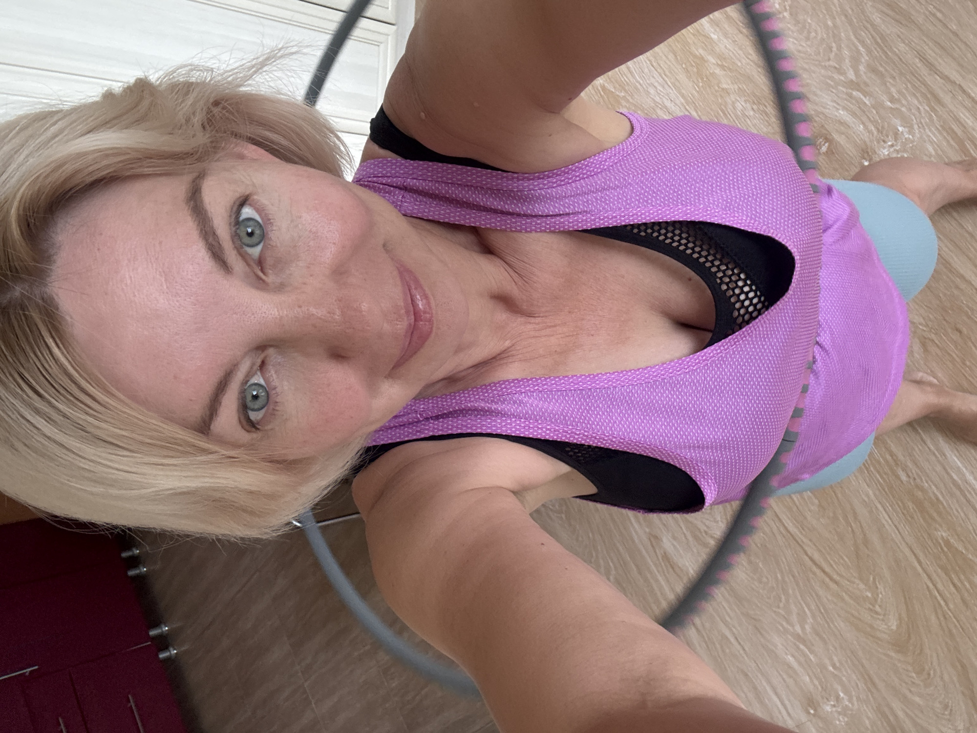 A woman with blonde hair and blue eyes is holding a hula hoop. She is wearing a purple tank top over a black sports bra and light blue shorts. She is standing on a wooden floor. The background shows a kitchen cabinet and a white wall. The image is a selfie taken from a high angle. The scene is an indoor setting. There is no information to suggest this scene is in Sochi, Russia.