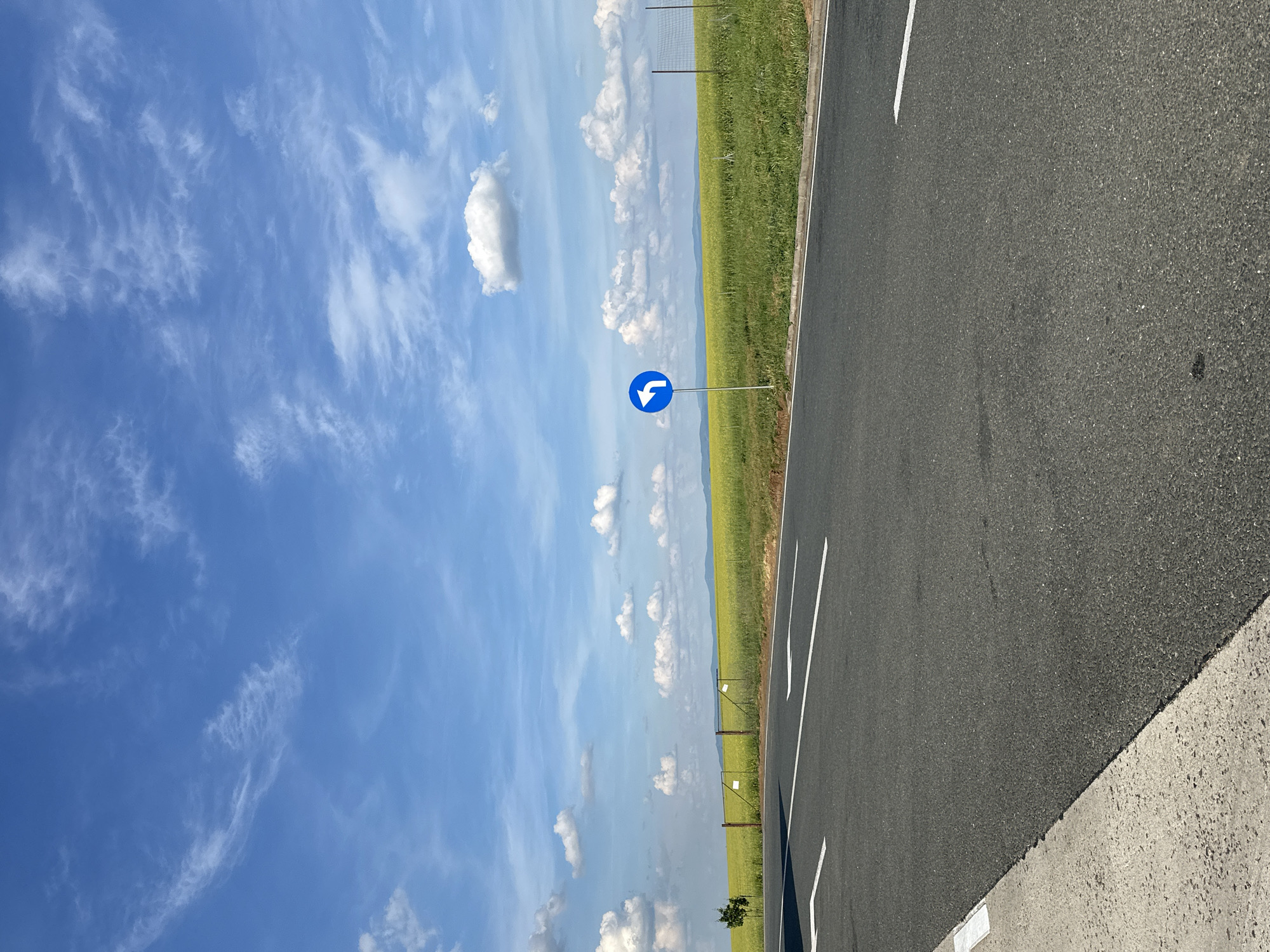 The image shows a road in Cruceni, Romania. The road is made of asphalt and has white lane markings. Next to the road is a field of green grass, enclosed by a fence. There is a blue road sign with a white arrow indicating a turn to the left. The sky is blue with scattered white clouds.