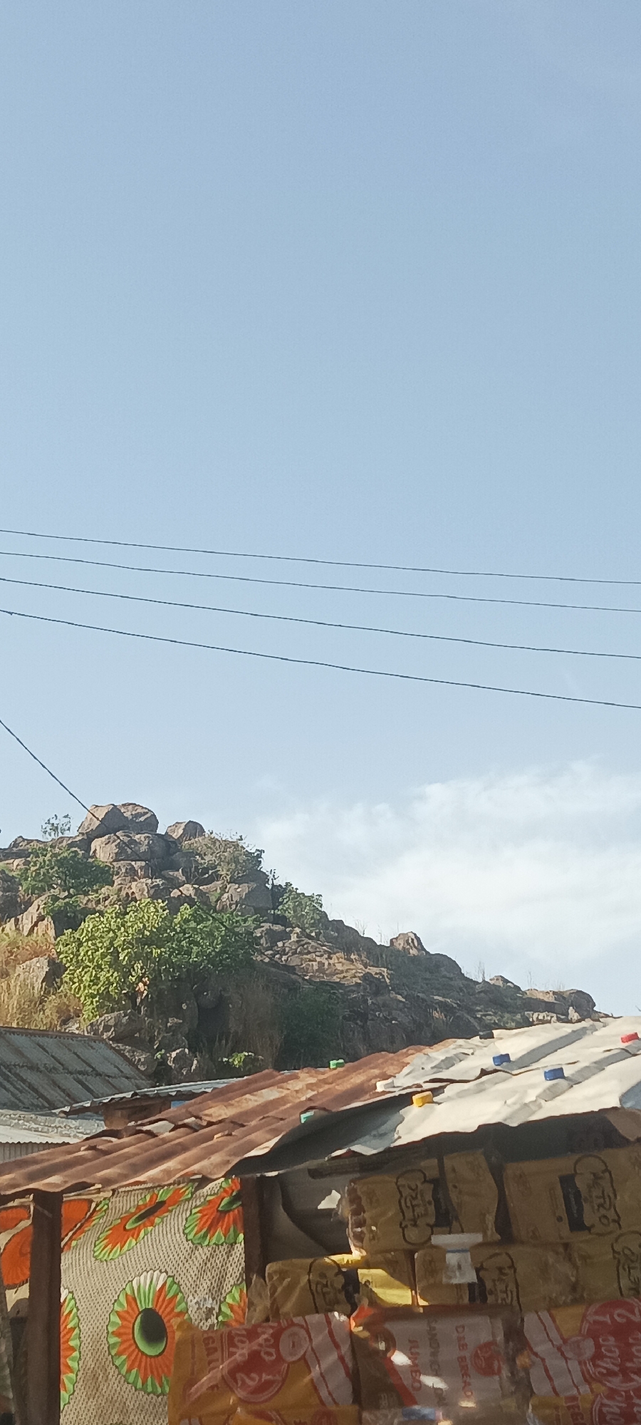 The image presents a bright, daytime view of a landscape in Kuru Dungus, Nigeria, encompassing natural elements and human structures. The upper half of the frame features a clear, light blue sky, with a few wispy white clouds visible towards the right. Several dark utility lines stretch horizontally across the sky.
Below the sky, a rugged, rocky hill dominates the mid-ground, characterized by large boulders and sparse green foliage interspersed with dry vegetation. In the foreground, the scene is anchored by the corrugated metal roofs of what appear to be small shops or market stalls. Some roofs show signs of rust, appearing brown, while others are a dull silver. Various colored plastic caps (blue, yellow, green) are scattered on these roofs.
On the left side of the foreground, a vibrant patterned fabric with orange and green circular designs is visible, likely forming part of a stall's covering. To the right, numerous packages of bread are stacked, indicating goods for sale. Visible text on these packages includes "METRO," "JUMBO," "TOP 2," "CHOP 2," "D&E BREAD," and "SAUSAGE BREAD." Some indistinct Arabic-like script is also visible on one package. The overall impression is that of a commercial area located near a natural rocky outcrop.