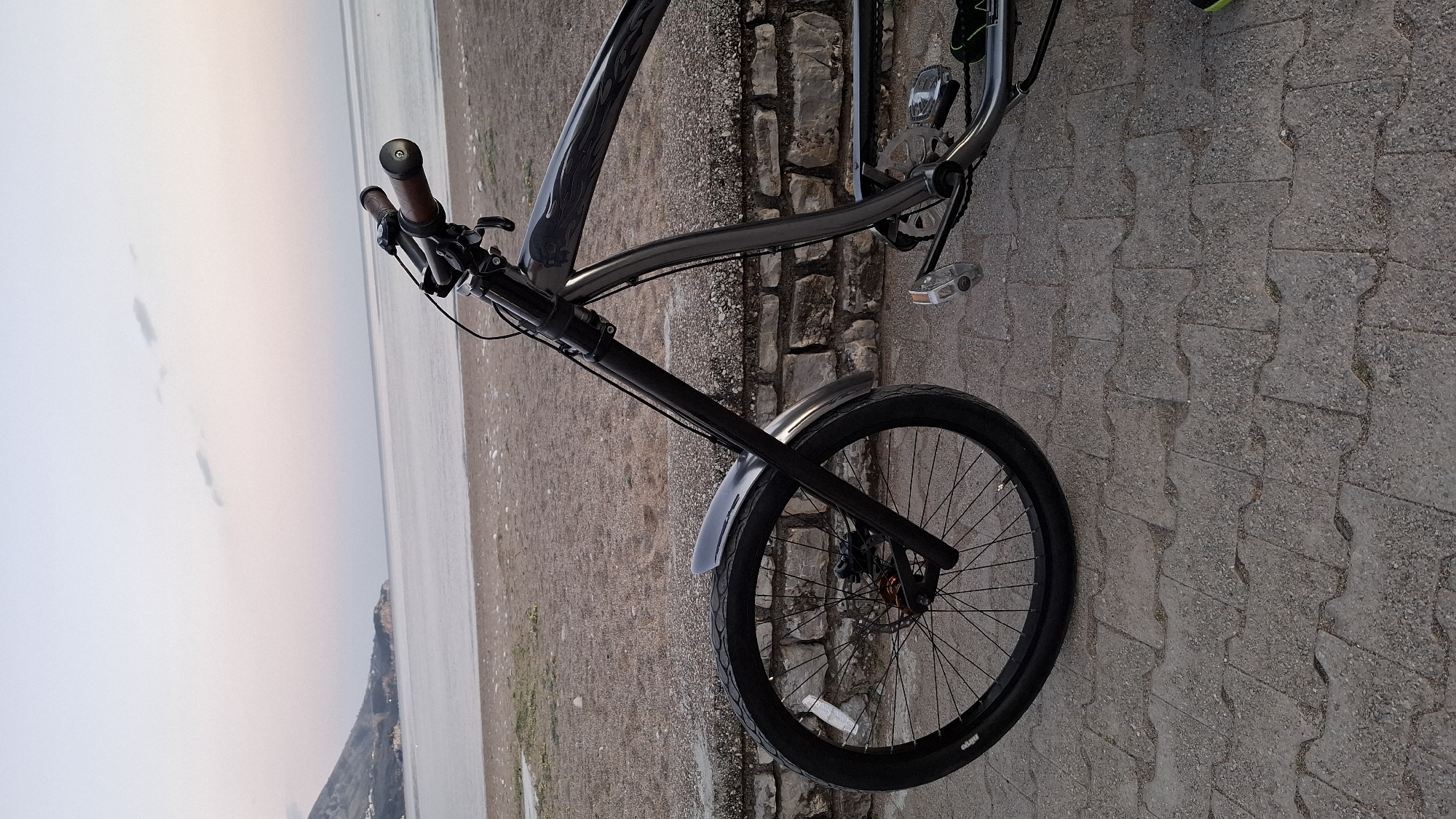 A bicycle is photographed on a cobblestone-like surface adjacent to a stone wall. The bicycle is leaning against the wall, with the front wheel prominently displayed. A body of water is visible in the background, potentially a bay or the sea, with land or mountains on the far side. The sky is overcast, creating soft, diffused lighting. The specific location is the city of Budva, Montenegro.