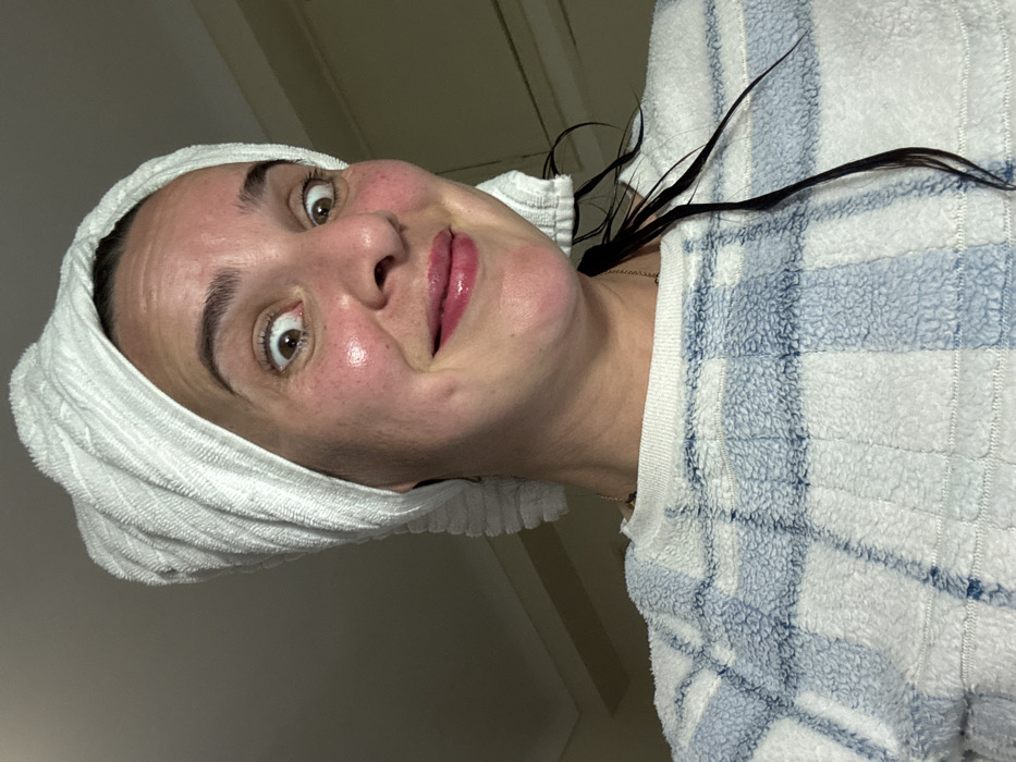 A woman with a white towel wrapped around her head is captured in a close-up, slightly rotated view. Her skin appears fresh, with visible pores and a slightly flushed complexion. Dark eyebrows frame wide, expressive brown eyes. She has a subtle, glossy lip color. Strands of dark hair peek out from under the towel and drape down her neck. She wears a soft, white and blue plaid top, and a gold chain is visible around her neck. The lighting is soft and diffused, creating a casual, candid atmosphere.