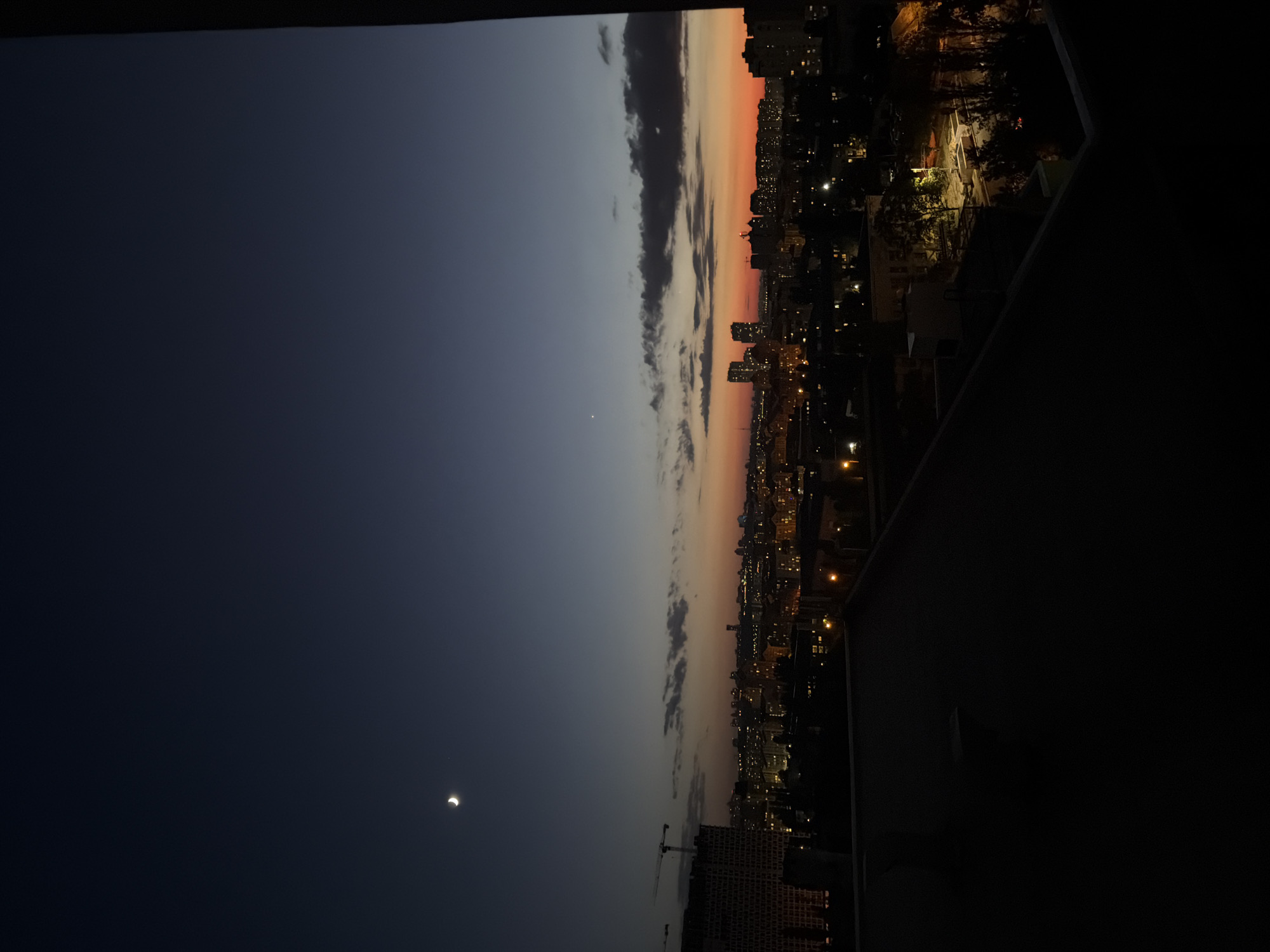 The image shows a twilight view of a city skyline, likely Kyiv, Ukraine, from a high vantage point. The sky transitions from a deep navy blue at the top to a warm orange and red hue near the horizon, indicating either dawn or dusk. A sliver of a crescent moon is visible in the upper left quadrant of the sky, accompanied by a faint star.
In the mid-ground, a cityscape is illuminated by a multitude of lights from buildings and streetlamps. Tall apartment buildings and industrial structures with cranes are visible, suggesting an active urban environment. The foreground is dominated by the dark silhouette of a rooftop or balcony, obscuring the immediate surroundings and creating a sense of depth. The overall atmosphere is serene and somewhat mysterious, capturing the transition from day to night.