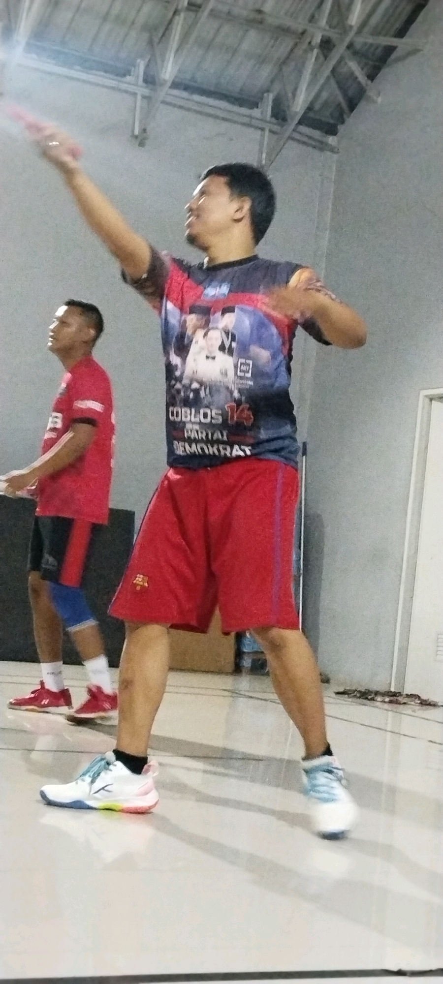 The image depicts two men indoors, possibly in a sports facility in Kliris, Indonesia, given the context. The foreground features a man wearing a dark t-shirt with a graphic print and red shorts. He is in motion, with his right arm extended upwards, holding a pink object, likely a badminton racket. His left arm is bent, and he appears to be mid-swing or preparing for a shot. He is wearing white and multi-colored athletic shoes. Behind him and to the left, another man is visible, wearing a red t-shirt, black shorts with red trim, and blue and black knee supports. He is also in a sporty stance, looking upwards. The environment appears to be a dimly lit indoor space with a light-colored ceiling structure and gray walls. The floor is light-colored and reflective. The graphic on the foreground man's shirt displays text that reads "COBLOS 14 PARTAI DEMOKRAT" along with images of people.