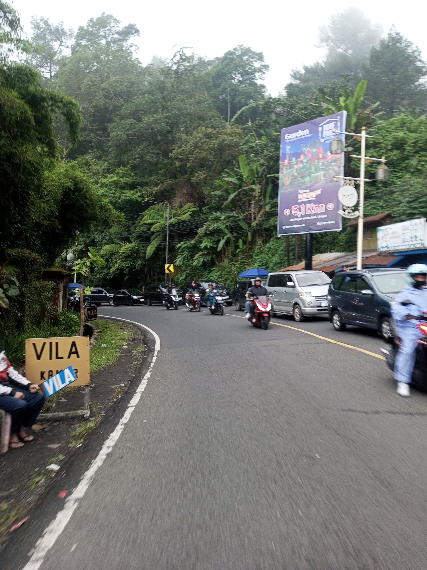 The image depicts a road scene in a tropical or subtropical environment. A paved road curves to the left, with a white line marking the edge. On the left side of the road, a person sits next to a sign that reads "VILA". The person is wearing a casual outfit consisting of what looks like a jacket and pants, and flip-flops on their feet. Behind the person, there is lush green vegetation, including bamboo.
Vehicles, including cars and motorbikes, occupy the road. There is an advertisement billboard on the right side of the road advertising a "Garden". The surrounding environment is marked by dense trees and foliage, with fog or low-lying clouds in the distance.