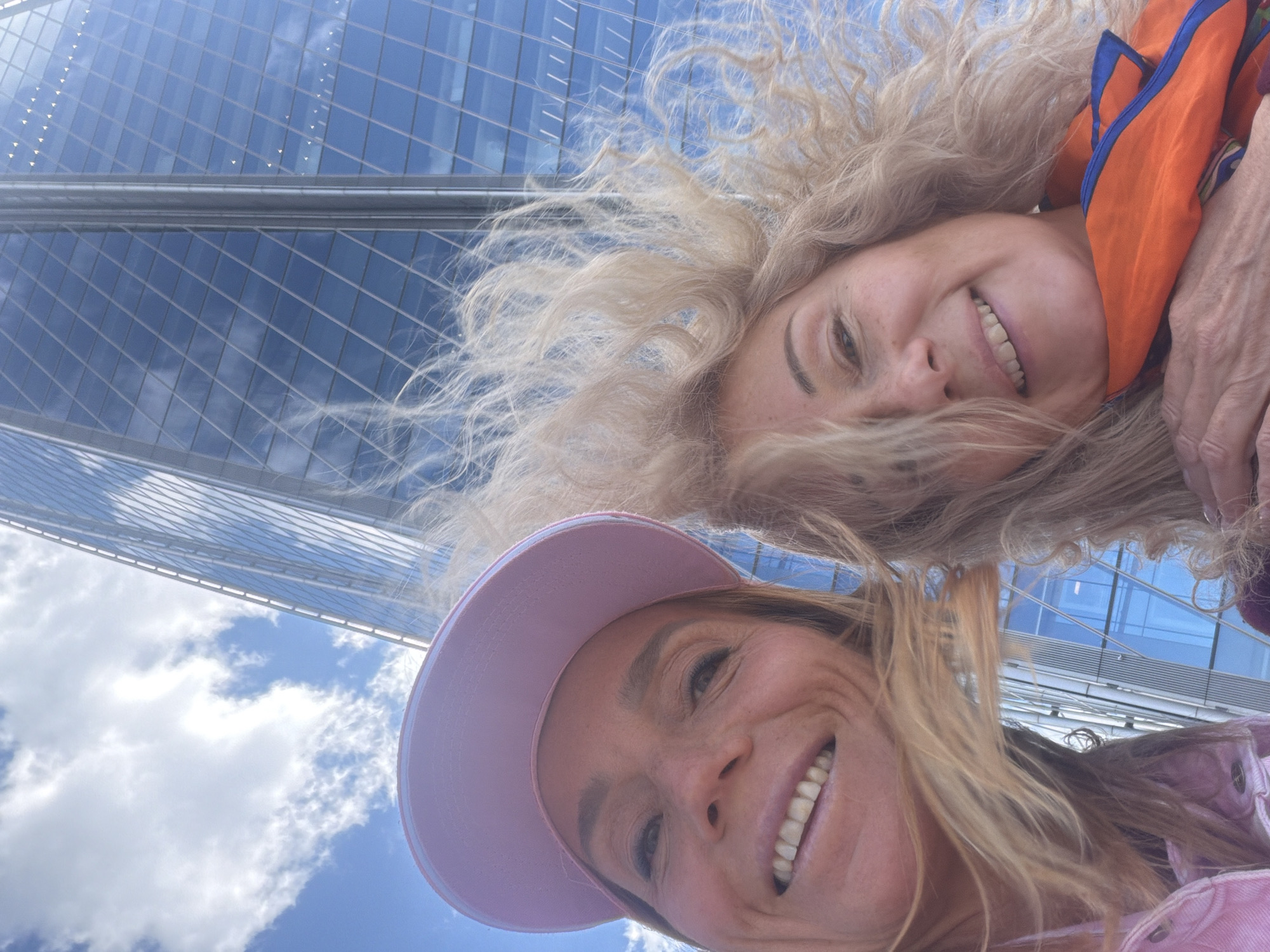 Two fair-skinned women are posing for a selfie in front of a tall, modern skyscraper with a glass facade reflecting the sky. The woman in the foreground is wearing a pink baseball cap and a light pink denim jacket, and she is smiling. The woman in the background has curly blonde hair and is wearing an orange and blue patterned scarf. The sky is blue with scattered white clouds. The location is likely Saint Petersburg, Russia, based on architectural styles and common landmarks.