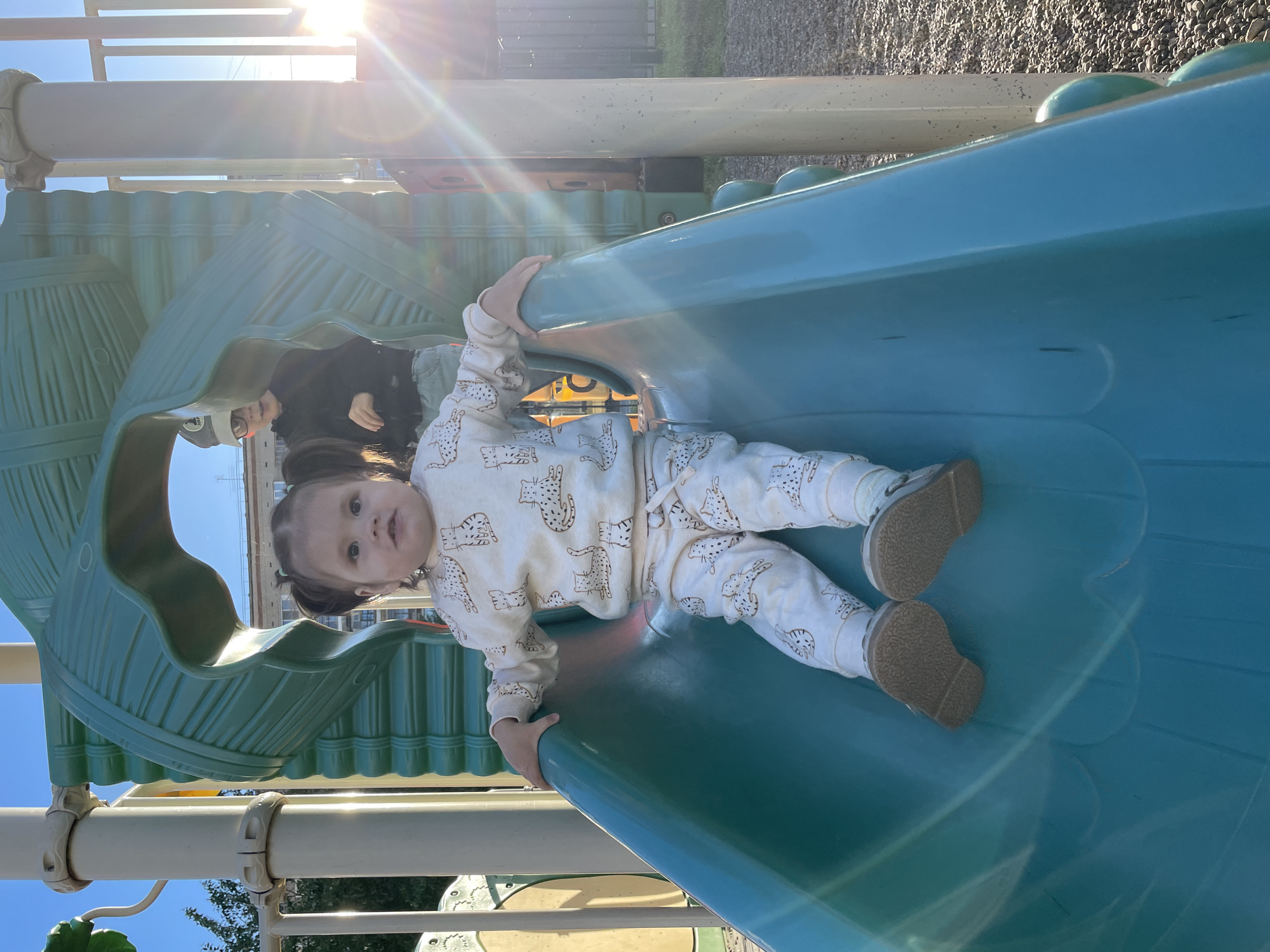A child wearing a white patterned pajama set and tan shoes is sitting on a blue playground slide. The child is looking directly at the camera. Behind the child, another person wearing a black shirt and light-colored jeans is sitting on a green part of the playground structure. The playground appears to be outdoors on a sunny day. The location is Izhevsk, Russia.
