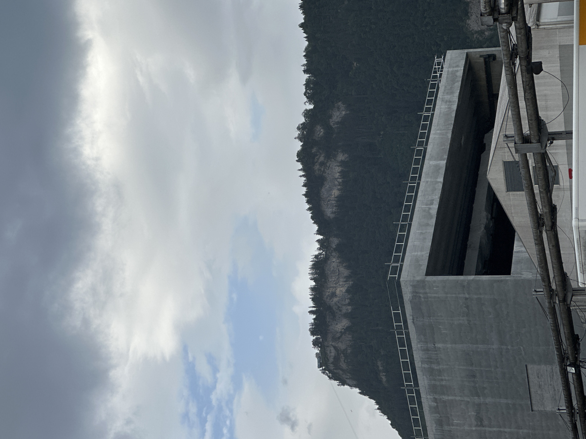 The image depicts a cloudy, overcast sky above a treed mountainside in Dornbirn, Austria. In the foreground, a large, angular, concrete structure with a metal railing along its upper edge dominates the lower half of the frame. Below this structure, industrial piping and part of a white building with a sloped roof are visible. The mountainside, covered in dense, dark green trees, rises steeply, with a lighter, rocky cliff face exposed near its crest. A building is visible on the top of the mountain, and a cable stretches diagonally across the sky from the mountain.