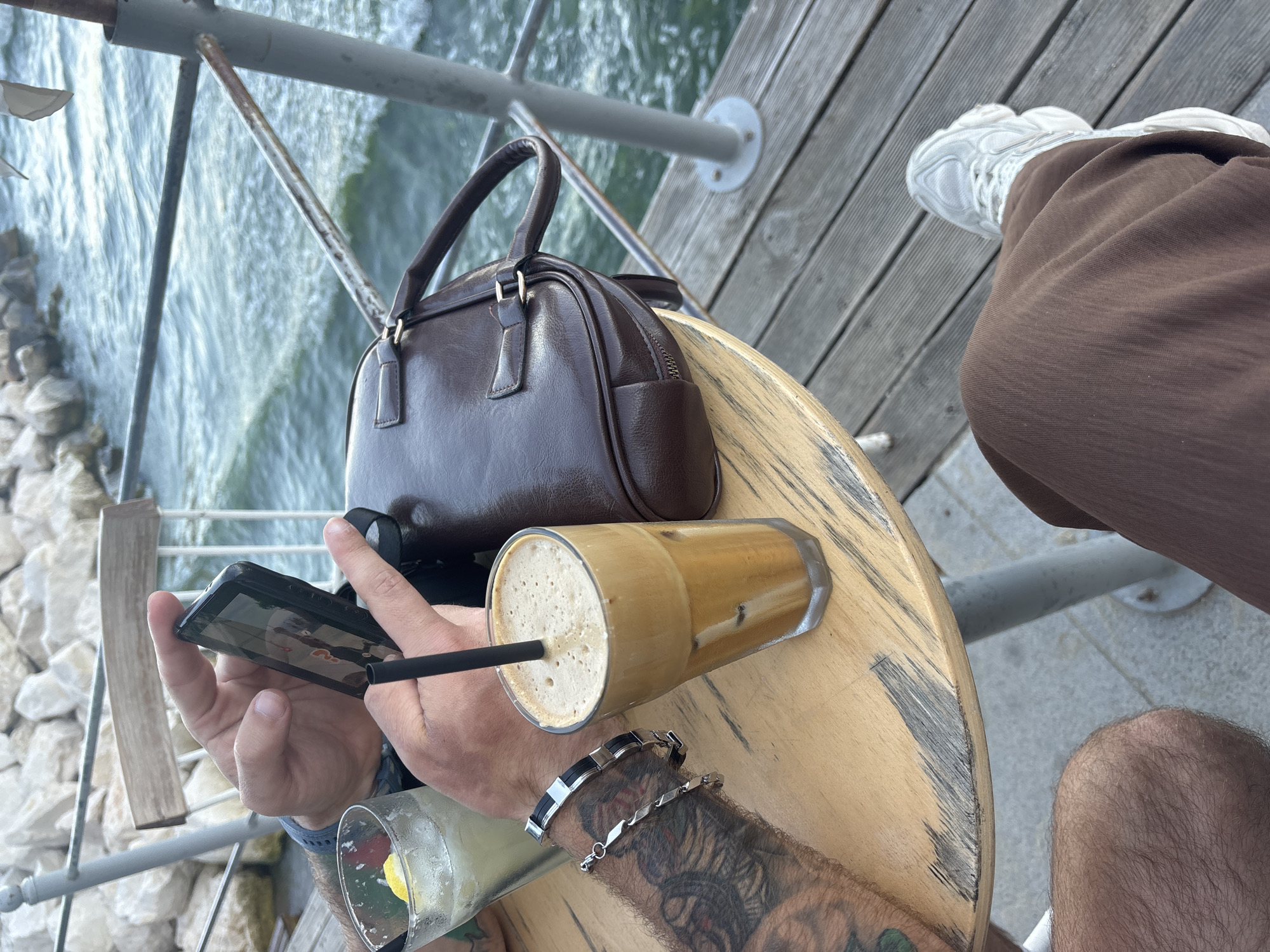This is a close-up, high-angle shot of a person's hands and a table setting outdoors, likely at a cafe or restaurant. The main subjects are a person's hands, holding a smartphone and resting on a wooden table, and several objects on the table.
In the foreground, a hand is holding a smartphone horizontally, with the screen displaying what appears to be a video or photo. The person has tattoos on their forearm and is wearing a silver bracelet with black accents and another delicate silver chain bracelet. To the right of the phone, a glass filled with a frothy, iced coffee drink is resting on the table, with a black straw inserted. Further to the left, another glass, partially visible, contains a lighter-colored drink with a slice of lemon.
Behind the drinks and hands, a dark brown, structured handbag with a zip closure and two top handles is placed on the table. The table itself is made of light-colored wood with a distressed, weathered finish, showing circular marks and scratches.
In the background, a body of water, possibly the sea or a lake, is visible, with a rocky shoreline to the far left. A metal railing or fence separates the seating area from the water. The person is wearing brown trousers and white athletic shoes.
The lighting suggests it is daytime, likely a bright and sunny day, given the outdoor setting and the light reflecting on the water. The overall mood appears relaxed and casual, as the person is engaged with their phone and enjoying drinks outdoors. The location cues (water, railing) and the context of Varna, Bulgaria, suggest a waterside establishment. There is no visible text in the image.