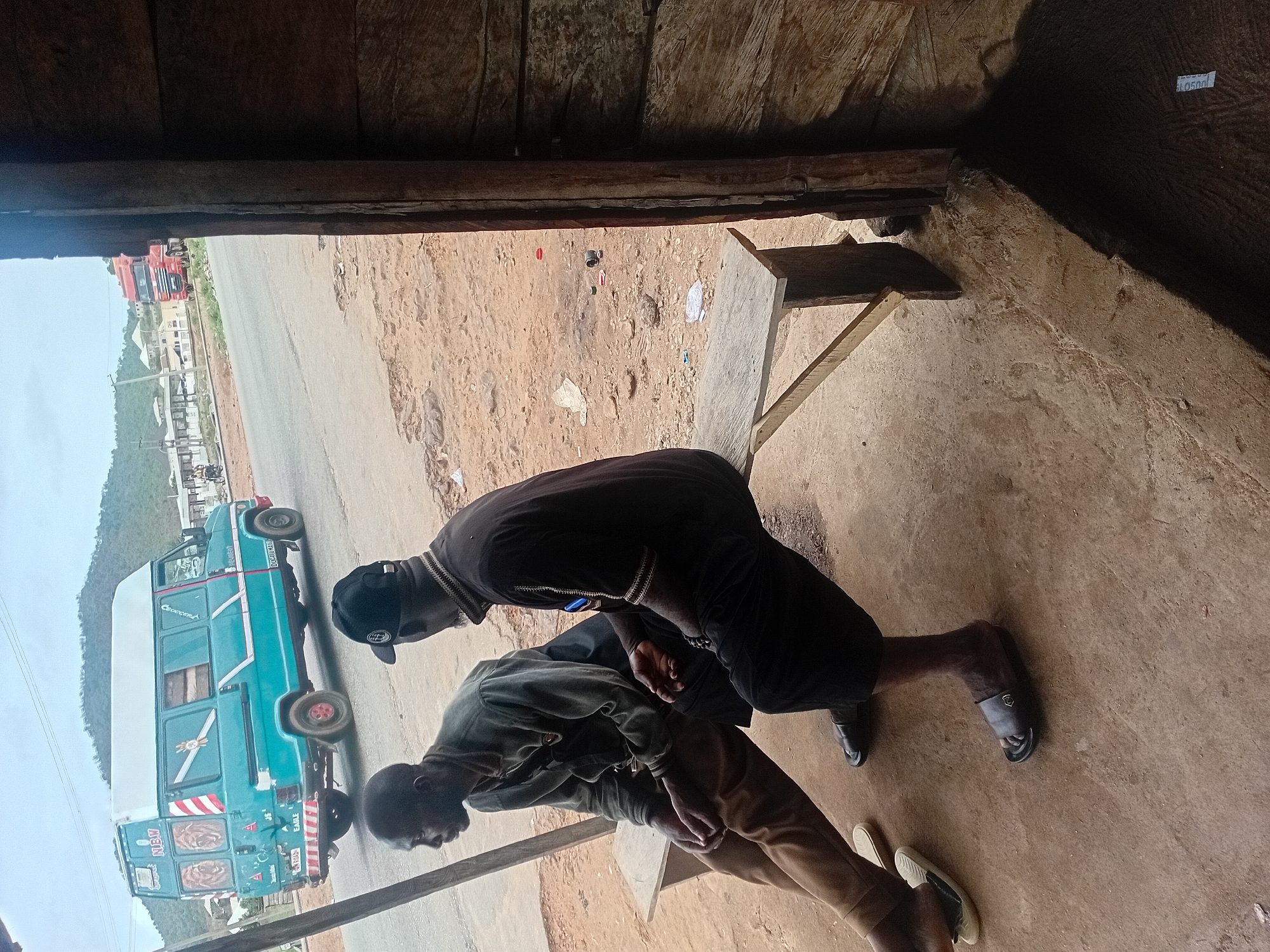Two men are standing in an enclosed wooden structure that opens onto a road. One man wears a dark shirt, dark shorts, a cap, and sandals. The other man wears a light-colored jacket, tan pants, and white sandals. A blue minibus is parked on the road outside the structure. A truck is also visible further down the road. The environment consists of a dirt road, some scattered trash, a building in the background, and a distant mountain. The location is in a rural area, in the city of Unknown, Unknown.