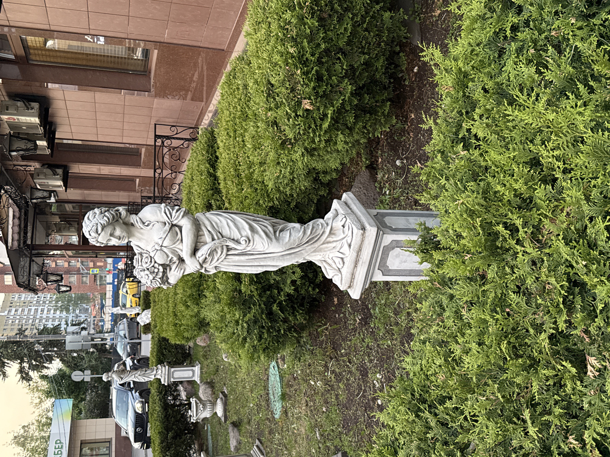 The image depicts a street scene in Moscow, Russia, featuring a classical-style statue of a woman with flowers, positioned among verdant shrubbery in front of a building with a marble facade. Further down the street, another, smaller statue of similar design is visible. Behind the shrubbery and statues, a road with vehicles, including a yellow taxi, can be seen. In the background, multi-story buildings line the street, and overhead power lines are visible. The environment suggests a blend of urban and landscaped elements within the city. A sign with Cyrillic script is visible on the left.