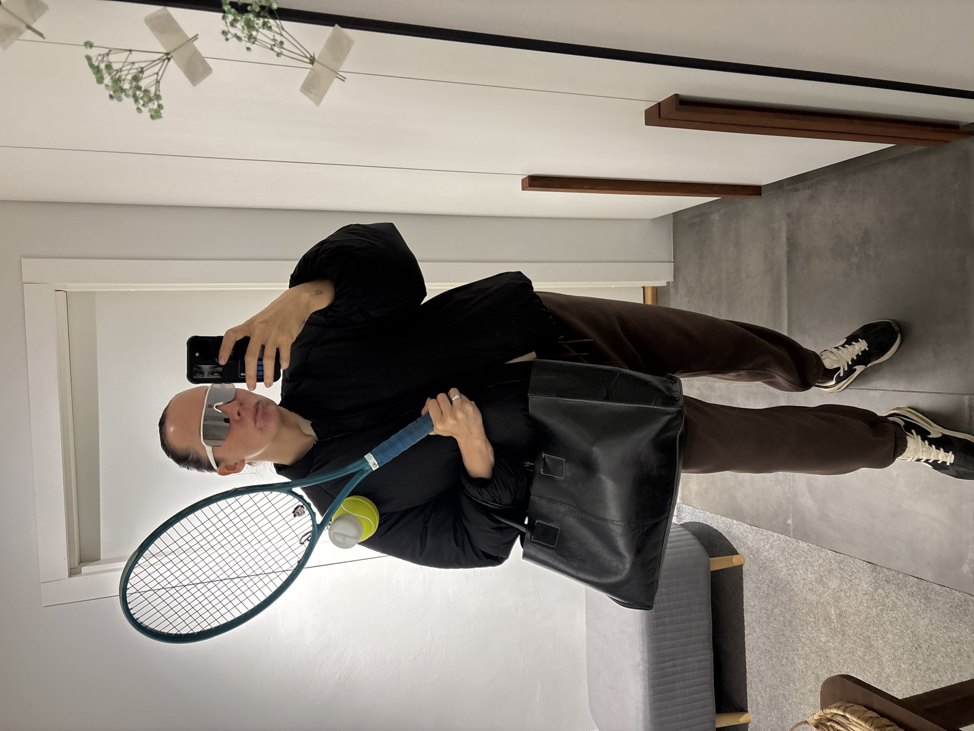 A person is taking a selfie in what appears to be an indoor space. The person is wearing brown pants, a black jacket, white-framed sunglasses, and black and white sneakers. They are holding a tennis racket with a yellow tennis ball and a black leather bag. The location is not possible to determine from the image.
