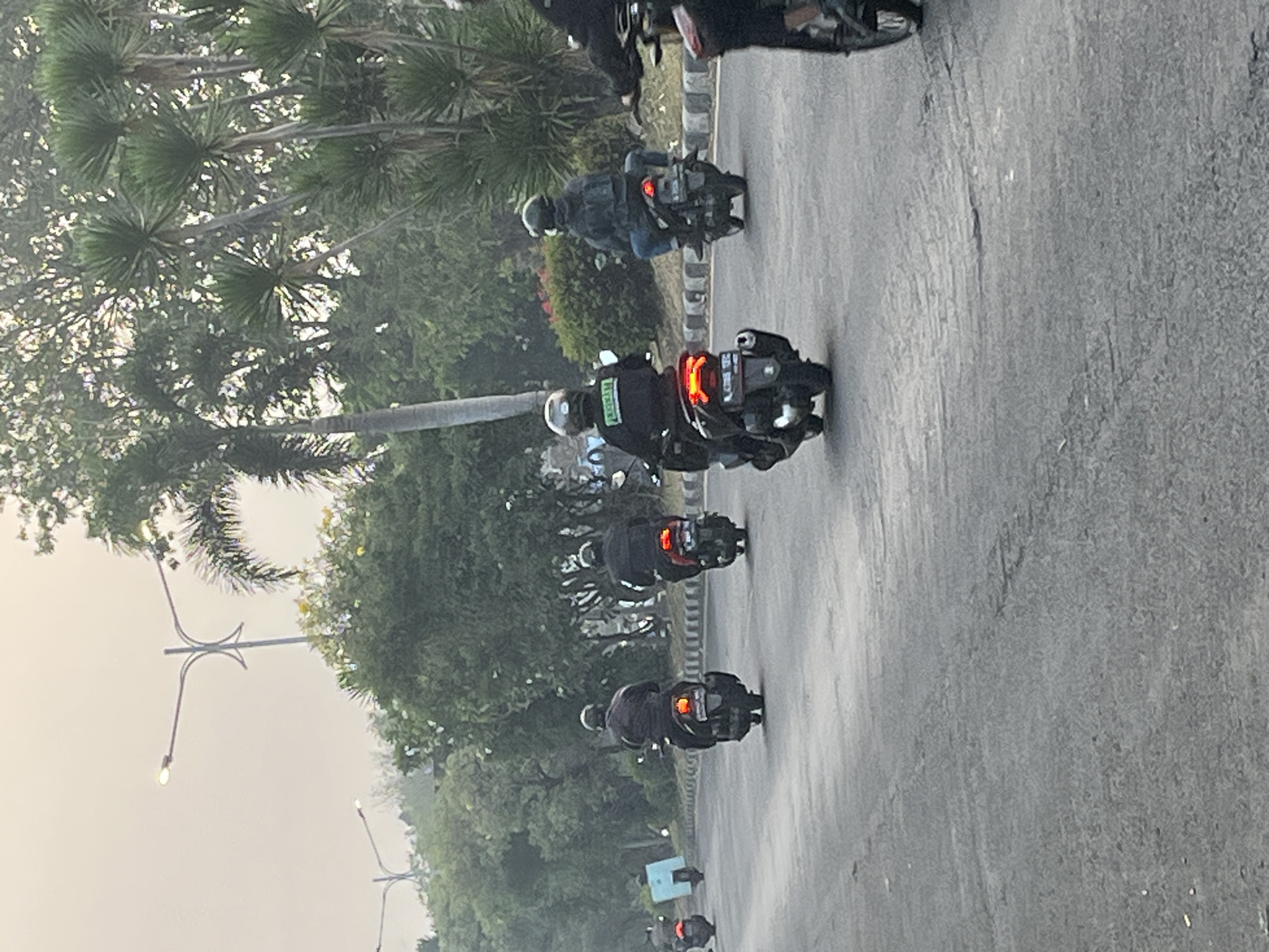 The image depicts a street scene in Kludan, Indonesia, on an overcast day. Several individuals are riding motorcycles on a paved road. The motorcycles are generally dark in color, and the riders are wearing helmets. One rider in the foreground wears a dark jacket with a green emblem. The road is lined with trees, including palm trees and other lush foliage, and a streetlamp is visible in the background. The overall impression is of a typical commute or travel in a semi-urban, tropical environment.