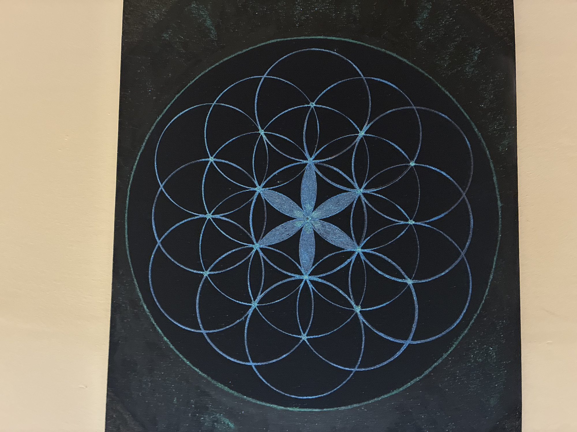 The image displays a black rectangular canvas featuring a geometric design known as the "Flower of Life." The design is rendered in shimmering blue and teal lines, creating a pattern of overlapping circles. The entire artwork is framed by a faint, circular outline in a green hue. The canvas is hung on a wall, with a pale, neutral-colored surface above it. The lighting suggests an indoor setting, with subtle texture and variations in the black background of the canvas. There are no people, animals, or other objects visible in the image. The image does not contain any information that would allow for its location to be identified in Alanya, Turkey, or any other city.