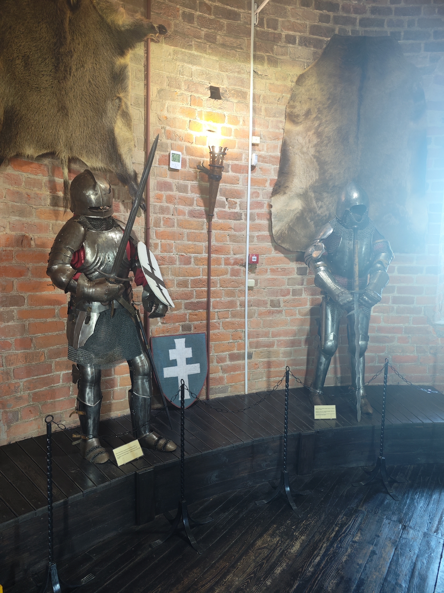 This is a full-shot, eye-level, interior photograph of two full suits of medieval armor displayed on wooden platforms in what appears to be a museum exhibit. The background consists of a red brick wall.
The suit of armor on the left is positioned slightly forward and to the left of the suit on the right. It is detailed with a helmet, gorget, breastplate, pauldrons, vambraces, gauntlets, a tasset, cuisses, and greaves. A chainmail skirt is visible beneath the breastplate, and a red tunic is worn underneath the armor. The knight holds a sword upright in its scabbard and a round shield with a white cross emblem is attached to its left arm. A white shield with a double cross emblem is propped against its left leg.
The suit of armor on the right stands further back and is positioned to the right of the other suit. It is also complete with all components of a full suit of armor. This knight holds a sword upright, resting the tip on the floor in front of its feet.
Both figures are standing on dark wooden platforms, which are separated by a black metal chain fence with black metal posts. The floor in front of the platforms is also dark wood.
Mounted on the brick wall above the left suit of armor is a large, taxidermied animal hide, possibly of a bear or wolf, with its head facing left. Above the right suit of armor is another, similar hide, but this one is largely obscured by a bright light source or lens flare emanating from the right side of the frame.
A torch sconce with a lit flame is mounted on the brick wall between the two suits of armor, casting a warm light. A digital thermometer is also mounted on the wall near the torch.
The scene is suggestive of a historical display, possibly within a castle or museum setting. This specific exhibit is located in the fortress of Kamyanyets, Belarus.