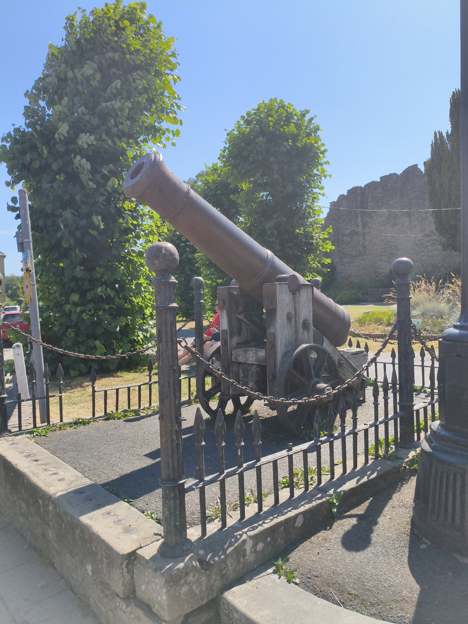 The image depicts a historic cannon, likely a relic from Ludlow Castle, positioned outdoors in what appears to be a public space within Ludlow, United Kingdom. The cannon rests on a wooden carriage with two large wheels. It is secured by a metal chain and is behind a decorative black metal fence with pointed finials. A stone wall, characteristic of historical fortifications, is visible in the background, as are lush green trees and a clear blue sky. The cannon is situated on a paved area with a low stone border in the foreground. A portion of a black lamppost is visible on the right side of the frame. Sunlight casts shadows on the ground, indicating it is a bright day. A person wearing a red shirt is seated on the carriage of the cannon.