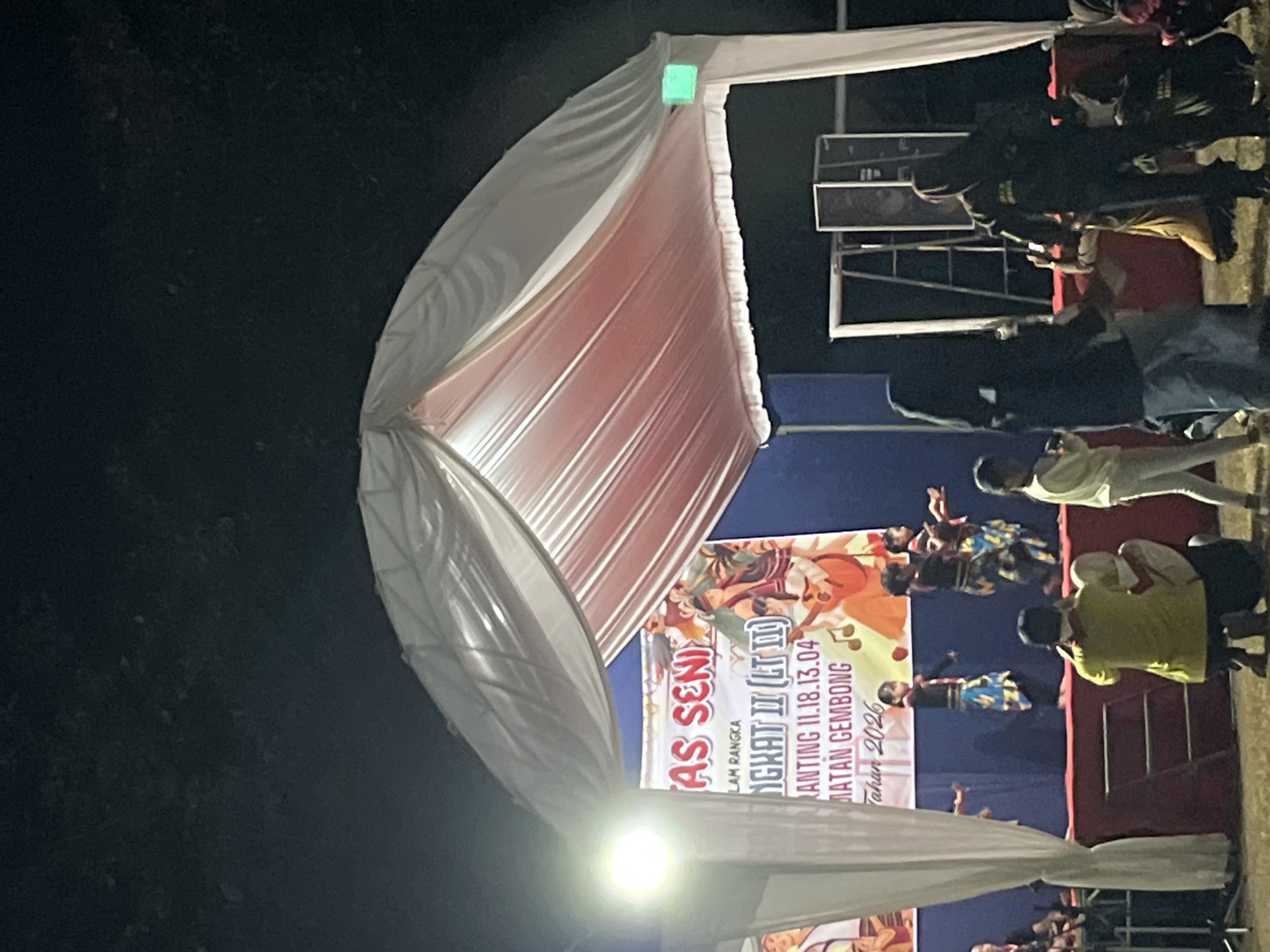 The image depicts an outdoor evening event in Gembong, Indonesia, featuring a stage set up under a large white tent with a pink interior lining. A banner on the stage reads "LAS SENI" and mentions "TINGKAT II (LT II) Gembong 11.18.13.04. Tataran Gembong Tahun 2026." Several children are performing a dance on the stage, dressed in traditional attire with colorful patterned skirts. In front of the stage, a crowd is gathered, some seated on the ground and others standing. A person wearing a yellow shirt is kneeling, seemingly taking photos. To the right, a group of people are also observing the performance. The environment appears to be a grassy field, with trees visible in the background, suggesting a rural or park-like setting within Gembong. The lighting is artificial, with a bright spotlight illuminating the stage and a green light attached to the tent structure. Two large speakers are positioned behind the stage.