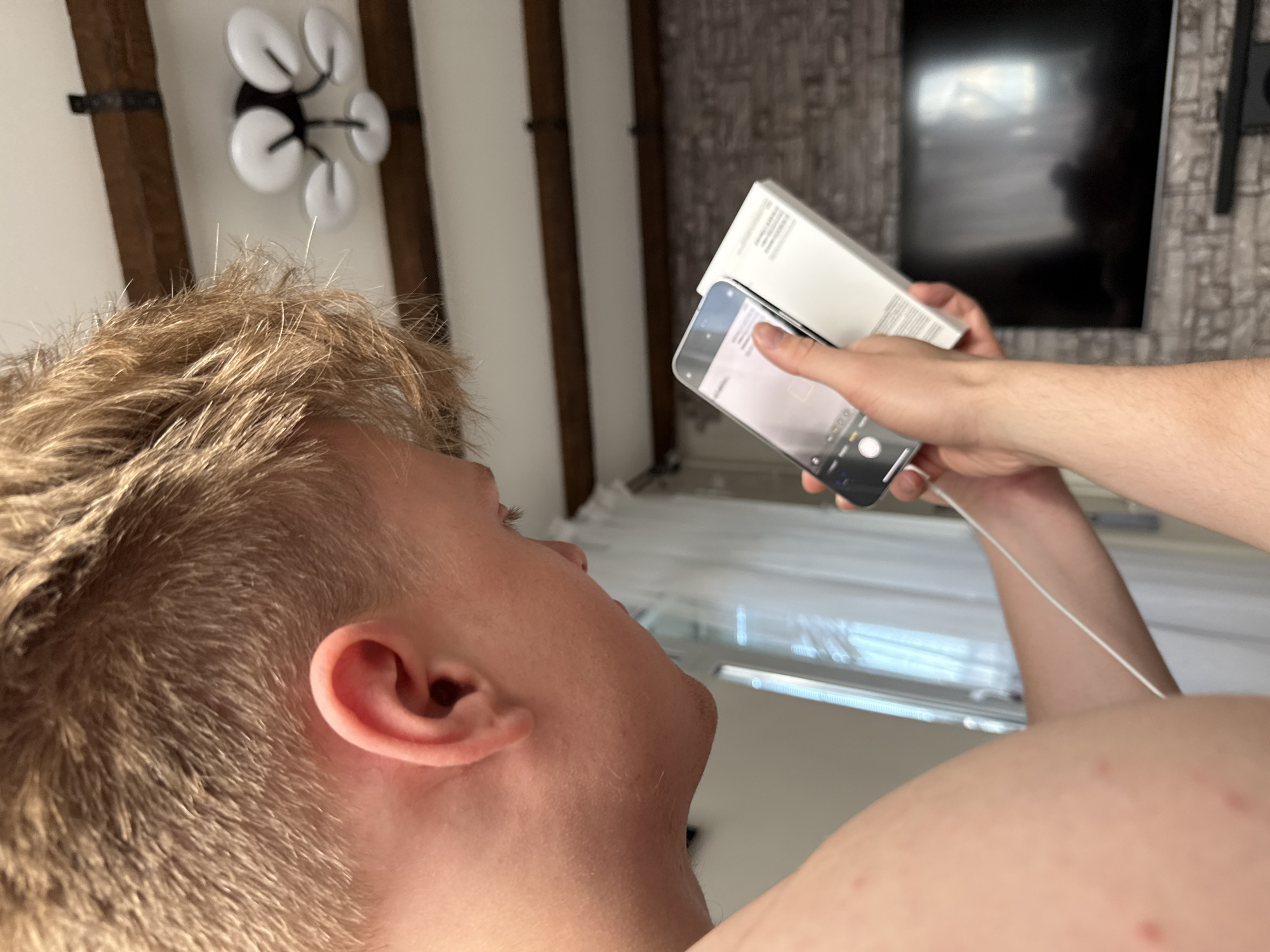 In the image, a person is holding a smartphone and a white box. The person's face and ear are visible in the foreground, with light brown hair. Their hand, holding the phone, is also prominent. The phone appears to be taking a photo or video of the white box, which is partially obscuring the background. The background includes what looks like a television screen mounted on a wall with a textured surface, and a ceiling with wooden beams and a light fixture. The overall setting appears to be indoors, possibly a living room. There is no discernible text in the visible parts of the image. The lighting suggests it could be daytime, but there are no strong indicators of the time of day or weather conditions. The activity is focused on the interaction between the person, their phone, and the box.