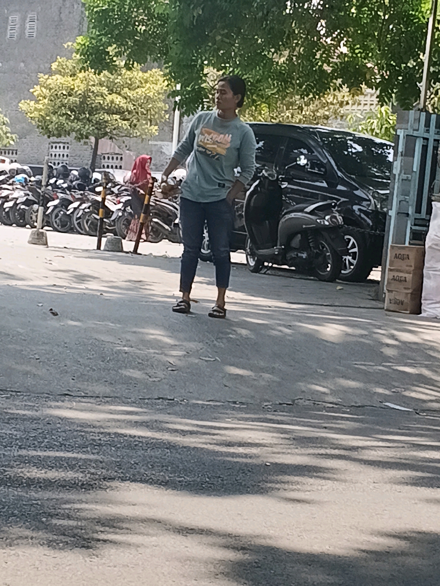 A person stands on a paved surface, with a row of parked motorcycles to their left and a parked black car behind them. The person is wearing a teal t-shirt with a graphic design and dark pants. They are looking to their right. In the background, there is a building with textured walls and trees. To the right of the frame, there are stacked boxes, one of which has the word "AQUA" printed on it. The scene appears to be an outdoor parking area in Purwodadi, Indonesia, indicated by the style of the motorcycles, the architecture of the building, and the presence of the "AQUA" brand, which is common in Indonesia. The lighting suggests it is daytime, with strong shadows cast on the ground.