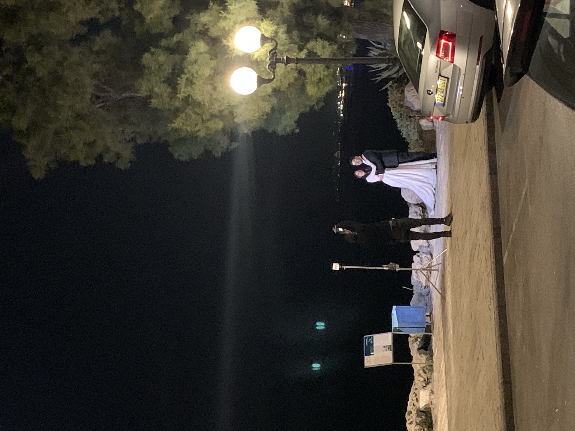 At night in Tel Aviv, Israel, a wedding is taking place outdoors. A bride and groom stand embracing near a lamppost. A photographer stands to the left of the couple, facing them. A car is parked on the right side of the image. In the background, there are lights reflecting on the water.