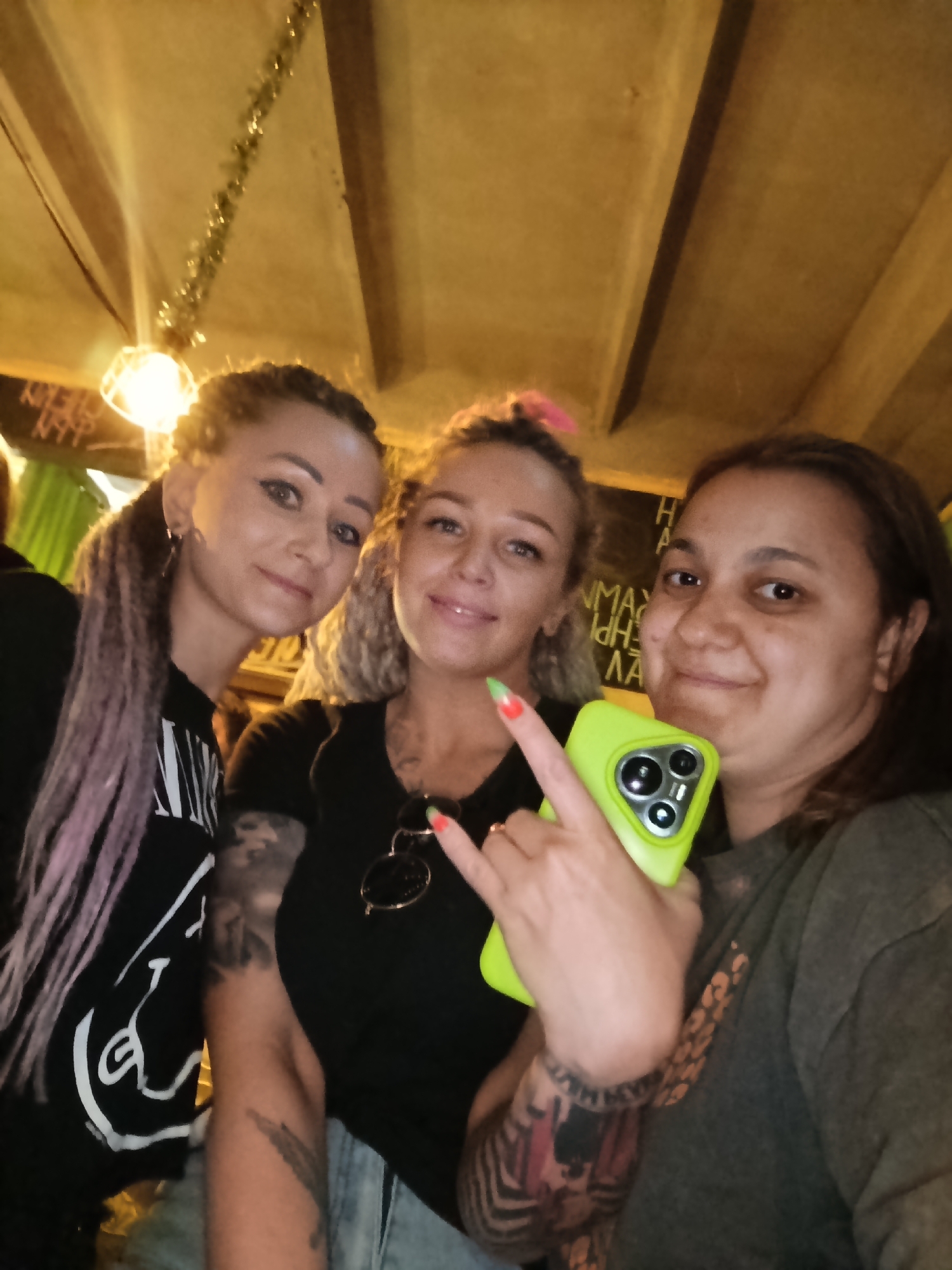 Three women are posing for a selfie indoors. The woman on the left has long, light purple braids and is wearing a black top with a white graphic. The woman in the center has blonde curly hair and is wearing a black top with sunglasses hanging off of it. She is making a rock-on hand gesture. The woman on the right is holding a bright green iPhone and has short dark hair. All three women have tattoos. The indoor setting has a wooden ceiling and a light fixture in the background. There is some writing on the wall behind them that is partially obscured. It is currently impossible to confirm the location of this picture is Chaikovsky, Russia.