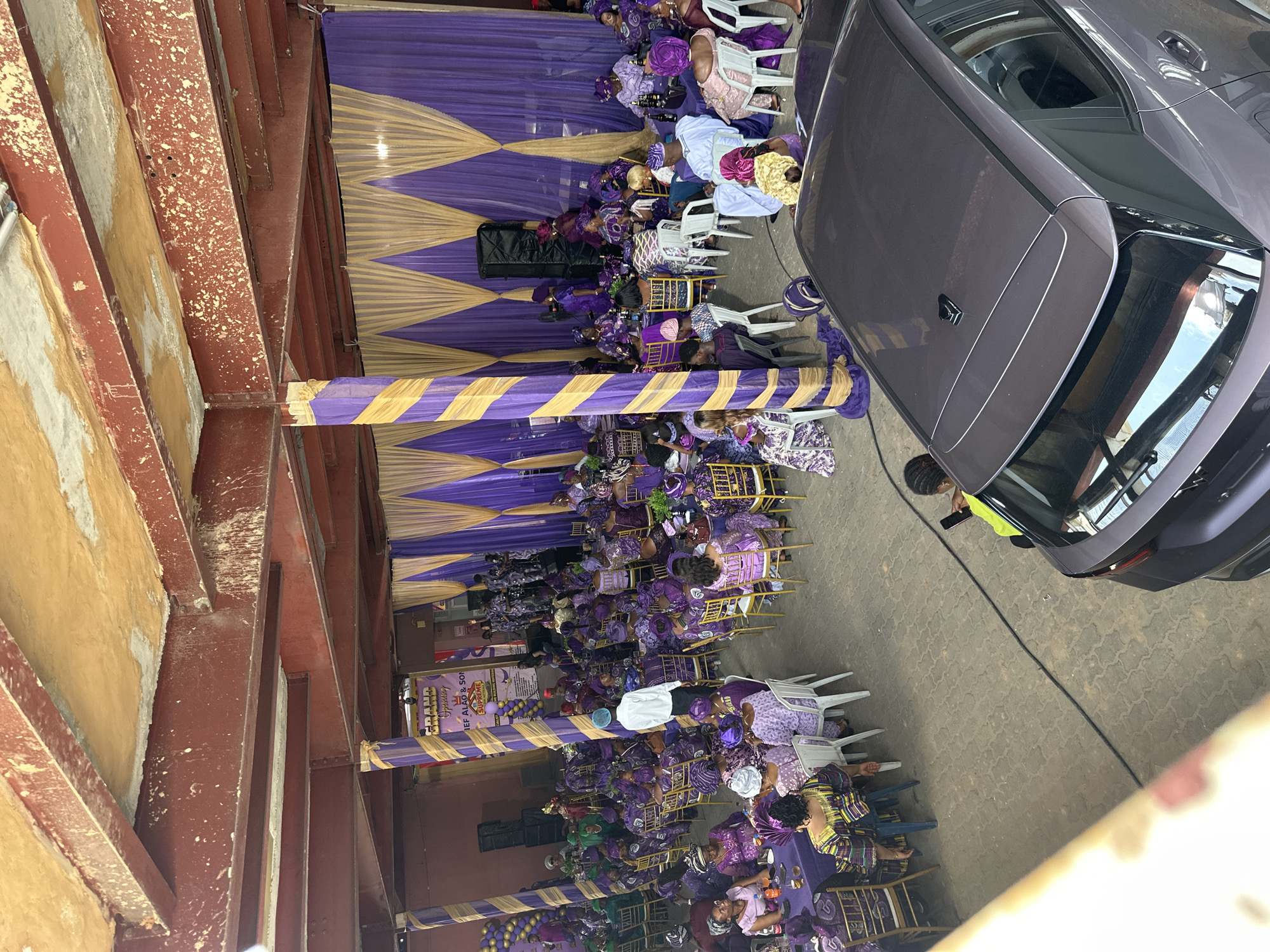 An outdoor event is occurring in Lagos, Nigeria, under a covered structure supported by exposed red metal beams. The event is decorated with purple and gold striped and geometric fabrics. Numerous people dressed predominantly in purple attire are seated in rows of white plastic chairs. Some are seated at tables covered with purple tablecloths. A large banner with text and images is present. A gray car is parked on the right side of the image. A person in a yellow vest is visible near the rear of the car.