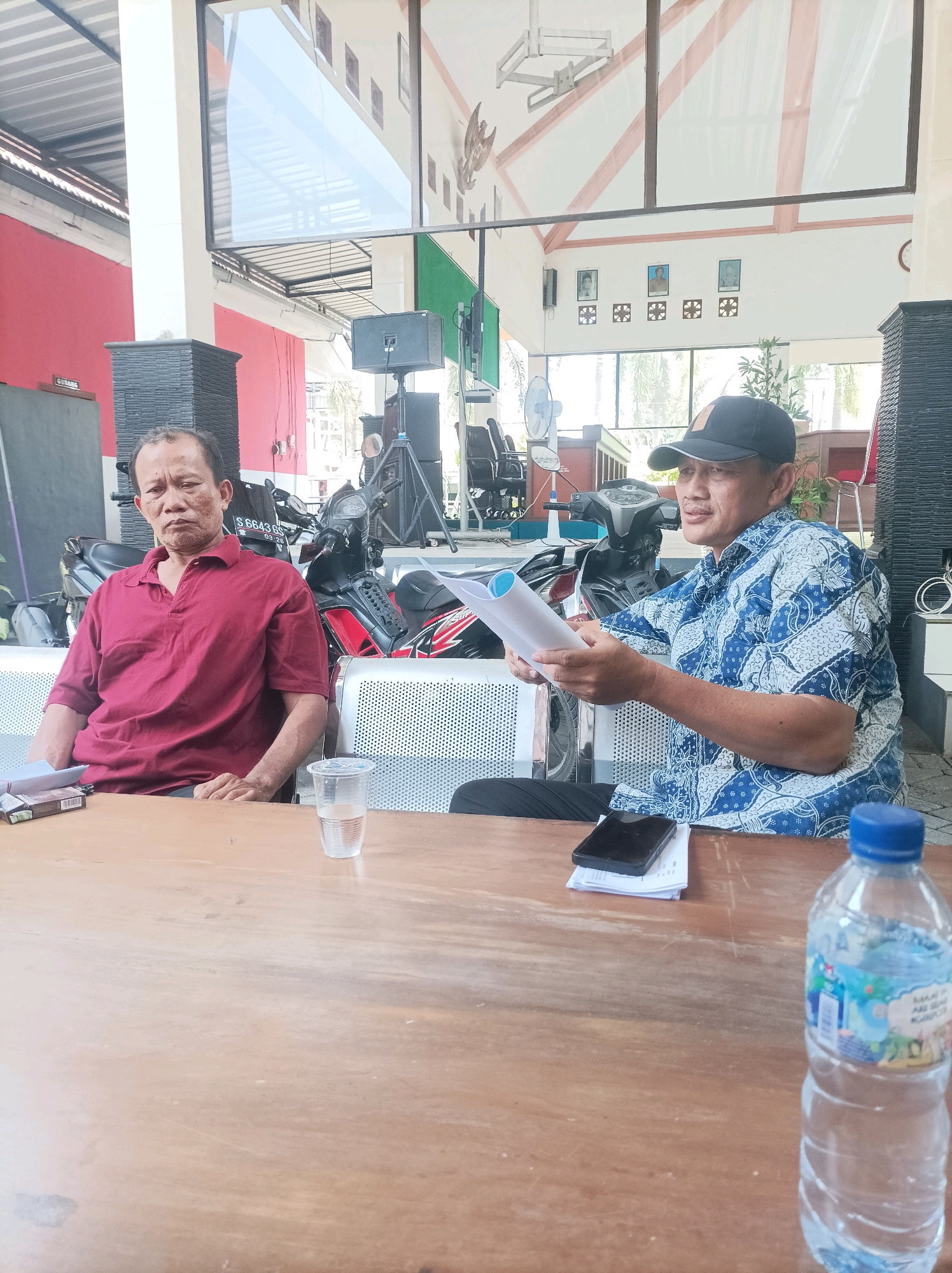 Two men are seated at a wooden table in what appears to be an outdoor covered area. The man on the left is wearing a maroon polo shirt and has his hands clasped on the table. The man on the right is wearing a blue batik-patterned shirt and a black baseball cap, and he is holding a few sheets of paper. A plastic cup of water, a pack of cigarettes, a smartphone, and a bottle of water are also on the table. In the background, there are motorcycles parked, and a building with a red wall and a white roof is visible. The overall environment suggests a casual meeting or discussion. The image includes Indonesian writing on a sign in the background, and the license plate on a motorcycle reads "S 6643 GS," which is a vehicle registration from East Java, Indonesia, indicating the likely location of Rengel, Indonesia, given previous information.